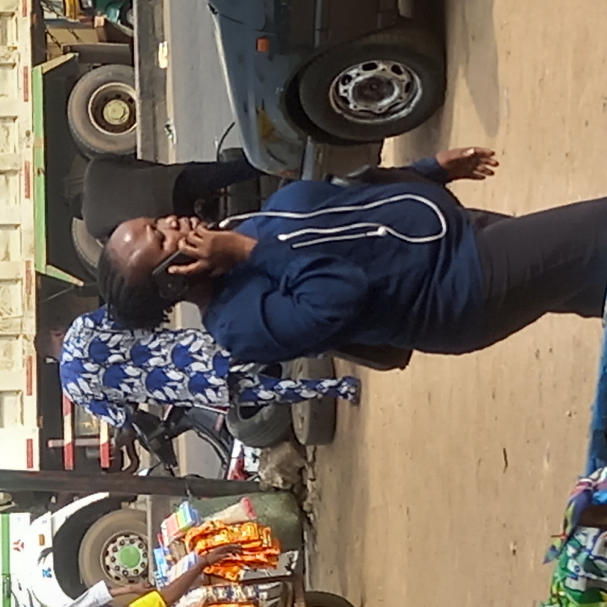 In Dadi Riba, Nigeria, a woman in a blue long-sleeved shirt and black pants is talking on a smartphone. She stands on a street next to a dark green vehicle and a large truck with a green stripe. Another person wearing a blue and white patterned shirt and pants is walking past her. A cart filled with packaged goods sits on the street near the bottom of the image. The ground is light brown and appears to be a paved surface.