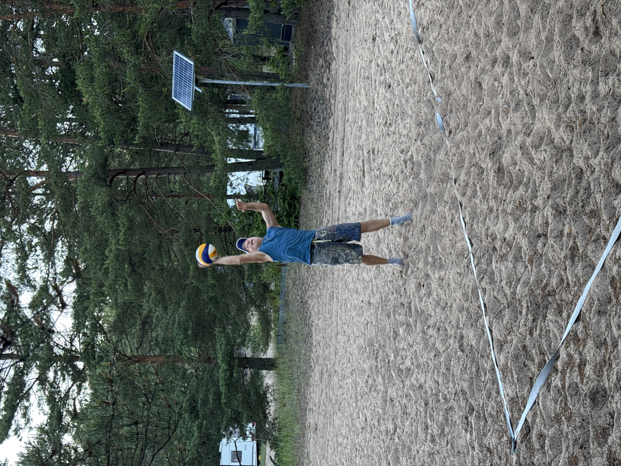 A man in a blue tank top and patterned shorts is playing volleyball on a sandy court. He is in mid-air, reaching up to hit the ball, which is above his head. He is wearing a blue baseball cap and grey socks. The volleyball court is marked with white tape on the sand. In the background, there are pine trees, a solar panel on a pole, and a recreational vehicle. The location is the city of Ozerki (Krasnodolinskaya), Russia.