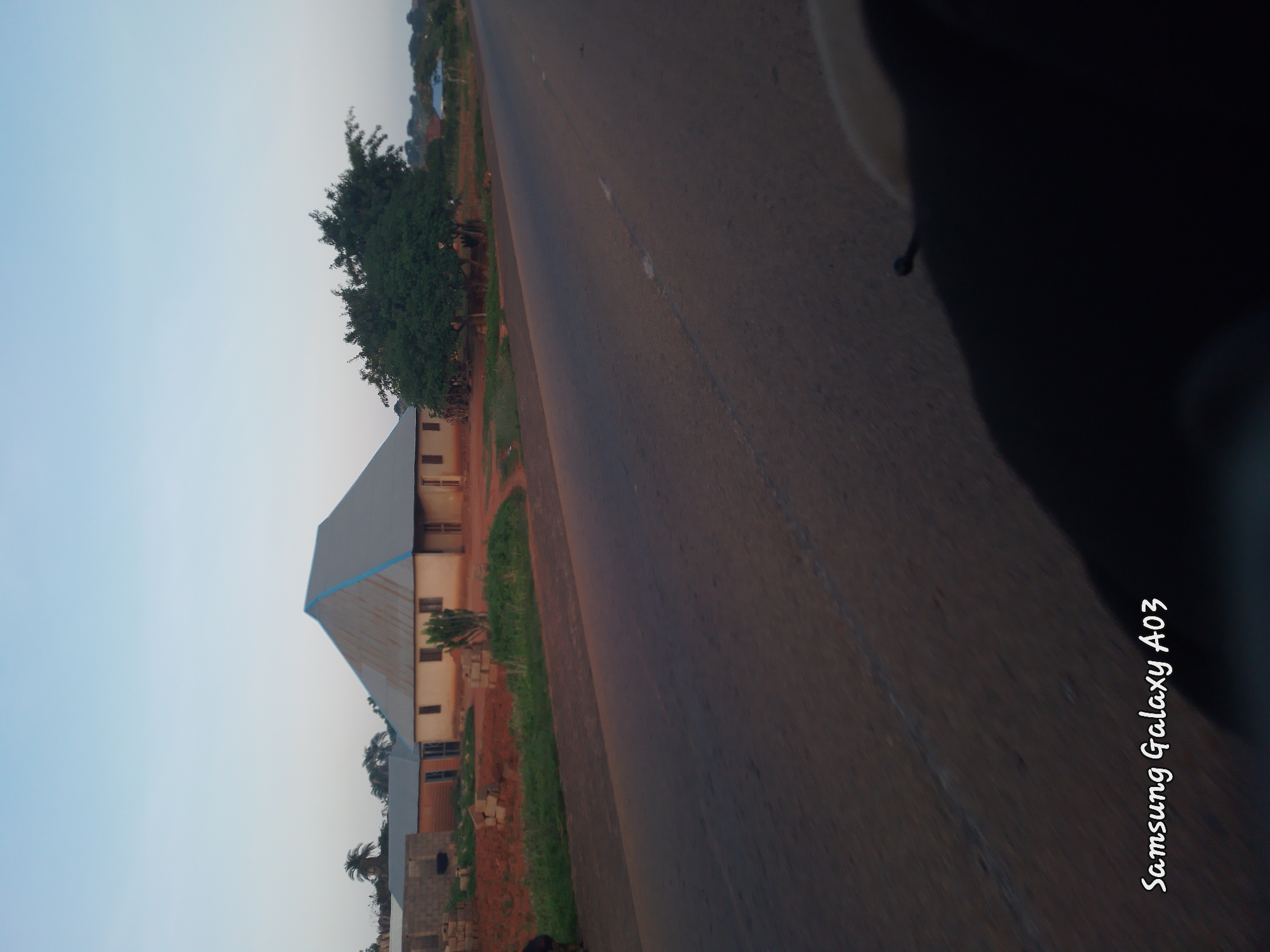 The image captures a roadside view in Doka-Tofa, Nigeria, under a clear, pale blue sky, likely during early morning or late evening. A paved road occupies the foreground, stretching towards the horizon with intermittent white markings. To the left of the road, a cluster of buildings is visible. The most prominent structure is a house with a large, pitched roof made of corrugated metal, painted in a light gray with blue trim along the edges. The house has visible windows and doors. Adjacent to it are other buildings, some appearing to be under construction with visible bricks. A few trees, including a larger, dense tree, are scattered around the buildings and the roadside. The ground appears to be red earth, typical of the region, with patches of dry grass. In the bottom right corner, a dark, out-of-focus object, possibly part of the vehicle from which the photo was taken, is visible. The bottom left corner of the image contains the watermark "Samsung Galaxy A03". No people are clearly discernible in the image.