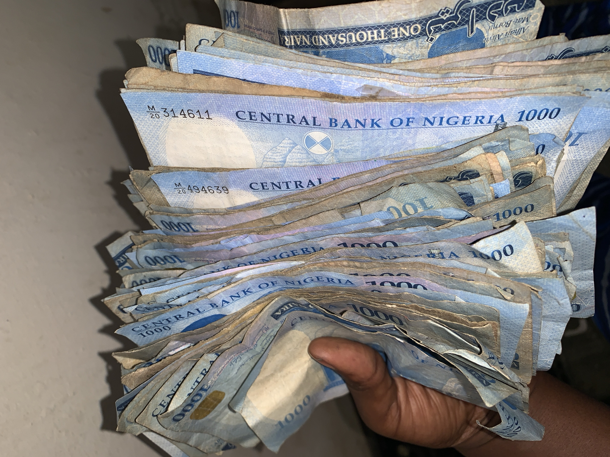 A hand holds a thick wad of Nigerian Naira banknotes, specifically the 1000 Naira denomination. The bills are stacked unevenly, showing signs of use, with some slightly worn edges. The banknotes display "CENTRAL BANK OF NIGERIA 1000" prominently. The serial numbers "314611" and "494639" are visible on two of the bills. Part of the top banknote reads "ONE THOUSAND NAIRA." The location, New Stadium in Nigeria, is not visually discernible from this image.