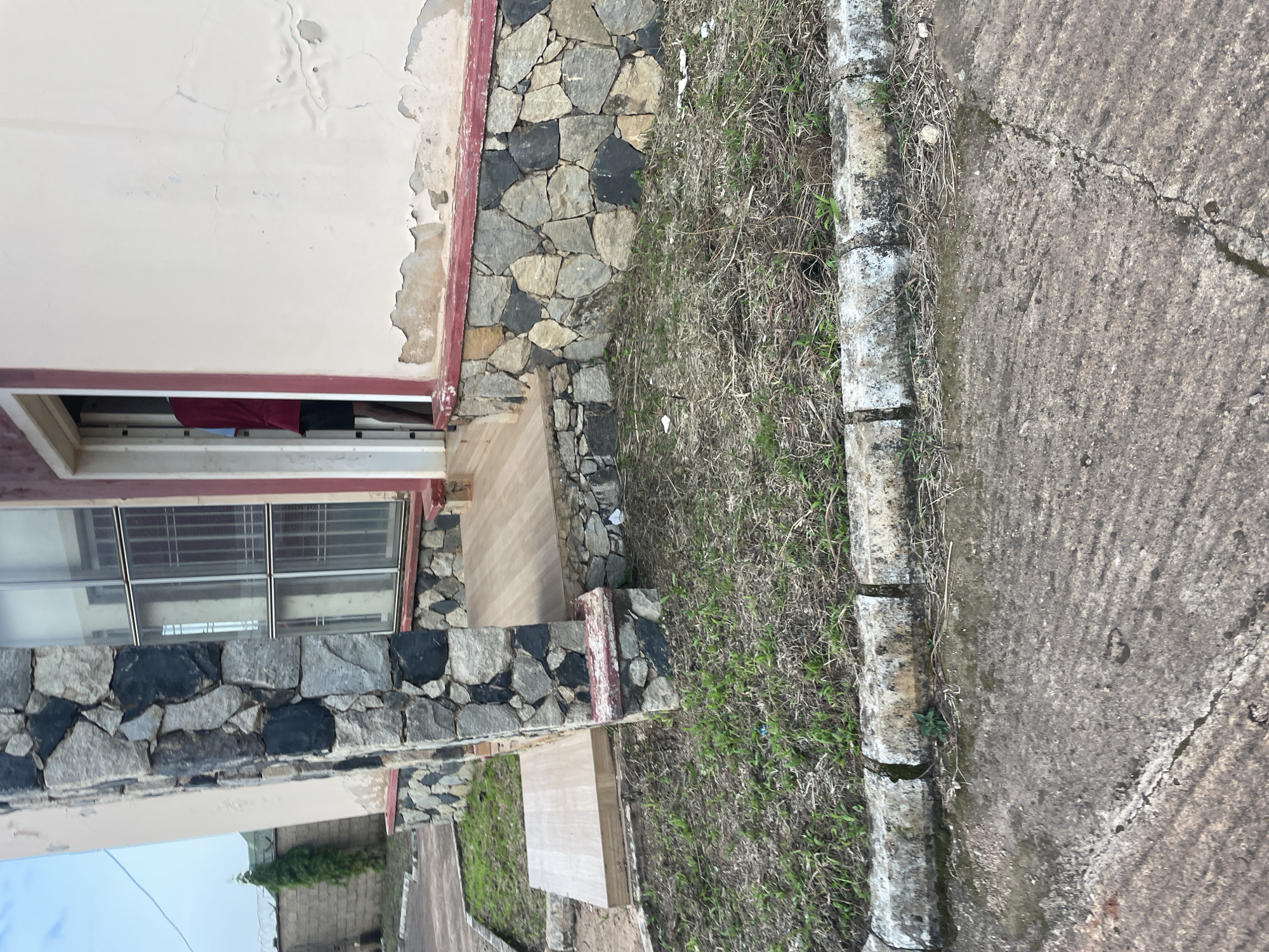 The image shows the exterior of a building in Jos, Nigeria. The building features a stone facade with a cream-colored upper section that shows signs of peeling paint. A doorway is visible, leading into the building, and a person wearing a maroon shirt and black shorts is partially visible inside. The ground in front of the building is covered with sparse grass and dry vegetation, bordered by a concrete curb. A paved area, possibly a walkway or road, is in the foreground, showing cracks and wear. The overall scene suggests a somewhat weathered urban environment.
