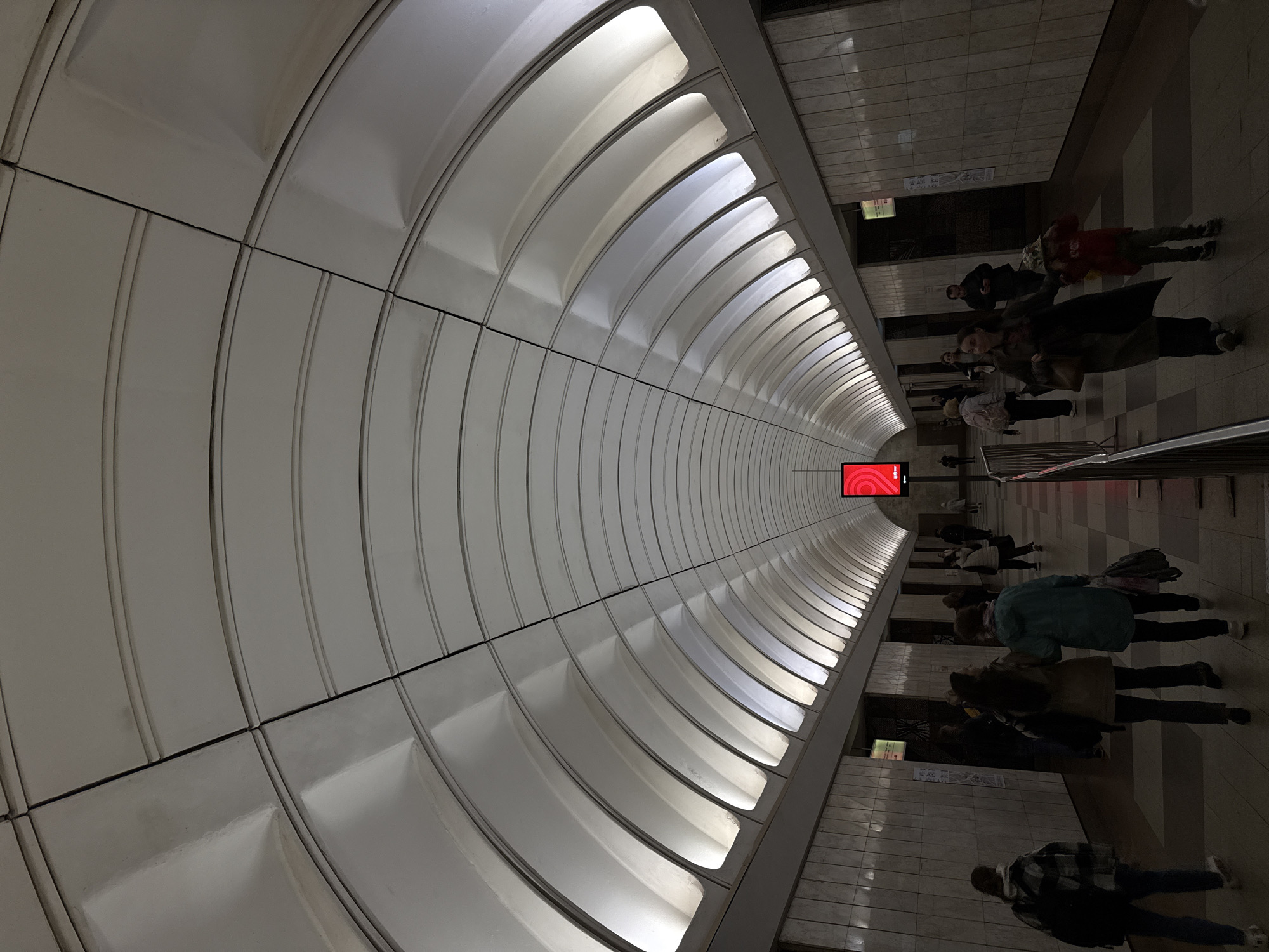 This media file depicts a view of a Moscow Metro station platform. The scene is characterized by the station's distinctive architecture, with a curved ceiling featuring arched illuminated panels. The walls are lined with white tiled sections.
Several people are visible on the platform, some standing and others appearing to walk. Their clothing suggests cooler weather. The lighting is a mix of the bright overhead illumination from the ceiling panels and the dimmer ambient light of the station, casting shadows on the tiled floor. The perspective of the shot is from a slightly elevated angle, looking down the length of the platform, which recedes into the distance.
In the distance, a red rectangular sign with a white logo or symbol is mounted on the wall. Further along the platform, there are tiled walls and what appear to be informational posters or signage. The overall impression is one of a functional, busy public transport hub. The time of day is indeterminate due to the artificial lighting.