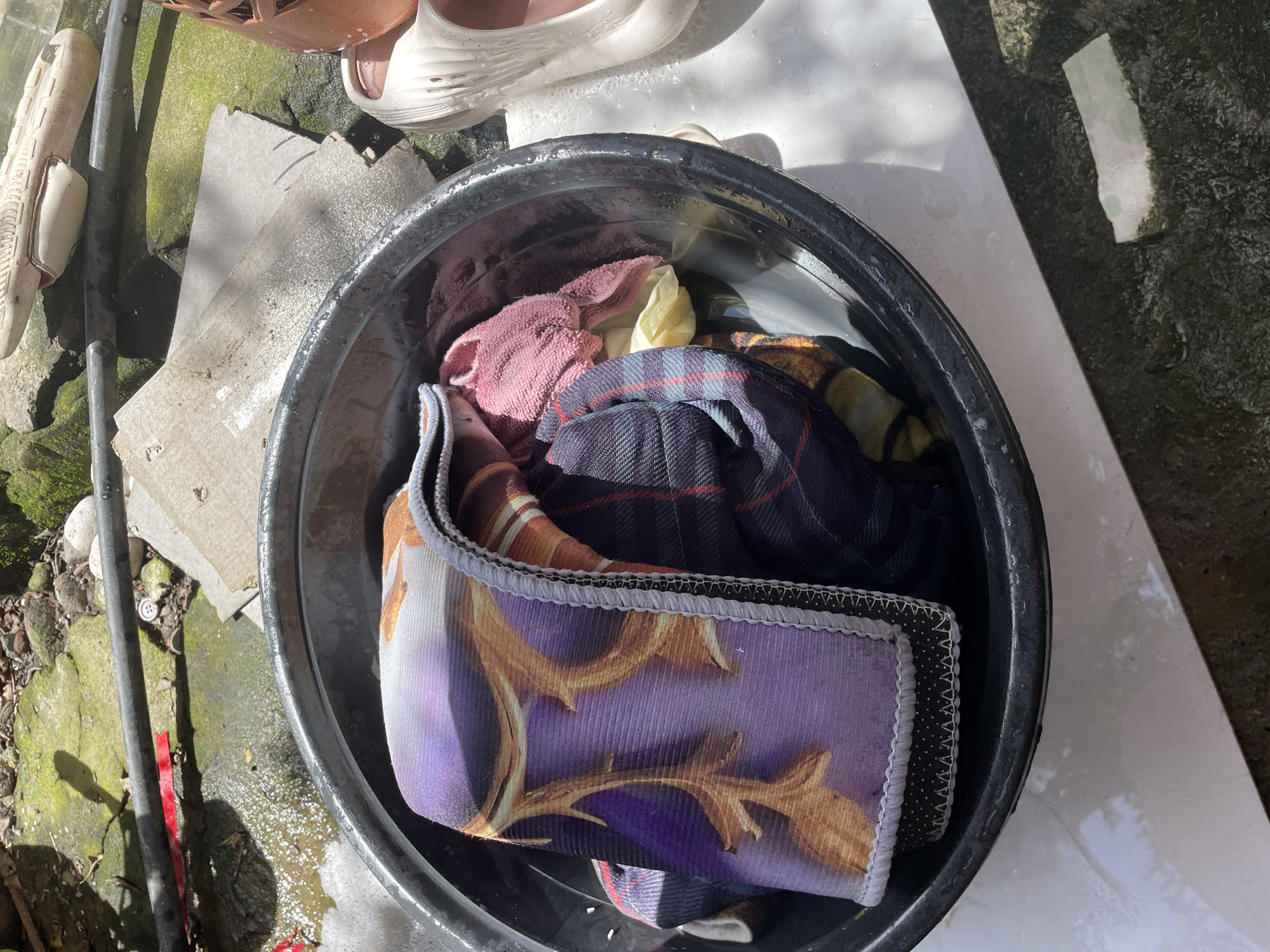 The image captures an outdoor scene centered around a black plastic basin filled with various items of clothing, suggesting laundry or soaking is taking place. Inside the basin, a prominent piece of fabric with a purple background and intricate gold or brown decorative patterns is visible, folded. Other textiles include a pink towel, a yellow cloth, and a dark blue or purple plaid fabric.
The basin rests on a ground composed of concrete, soil, and moss-covered areas. Bright, direct sunlight illuminates the scene, casting sharp shadows from the basin and other objects, indicating it is daytime. A pair of beige or white sandals, resembling Crocs, are partially visible next to the basin, along with a brown woven item.
Surrounding the basin on the ground are several pieces of light grey paper or cardboard, a black metal pipe, and scattered small rocks. A small white button is also discernible on the mossy ground. There are visible traces of water or wetness on the rim of the basin and on the ground nearby. The utilitarian outdoor setting is consistent with a household chore being performed in a sunny environment, such as in Davao City, Philippines. There is no visible text in the image.