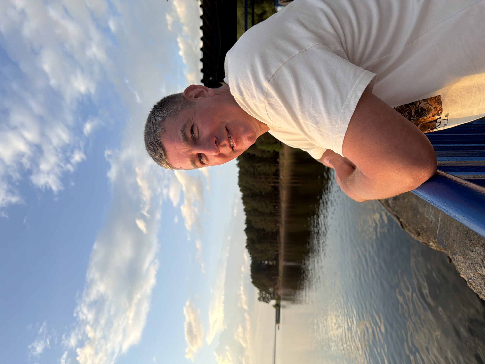 A man stands on what appears to be a pier or embankment in Chaikovsky, Russia, leaning against a blue railing. He is wearing a white t-shirt. Behind him, the skyline features trees and a body of water, possibly a lake or river, reflecting the sky. The sky has clouds. The photograph is oriented with the sky on the left and the water on the right.