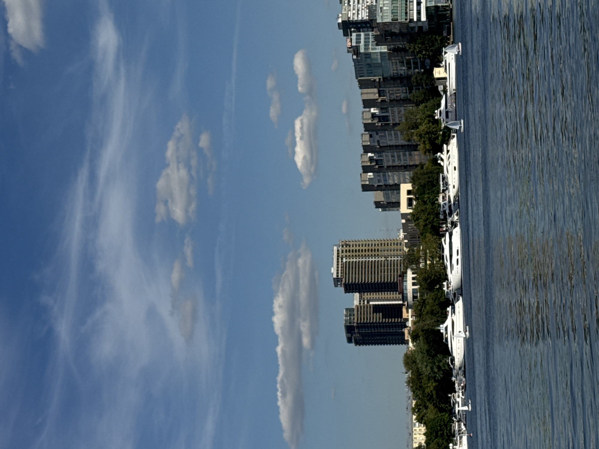 A wide-angle, eye-level shot captures a serene cityscape along a body of water. The sky is a vibrant blue, dotted with wispy white clouds. In the mid-ground, a collection of modern high-rise buildings forms the city's skyline, varying in height and architectural style. Lush green trees line the waterfront, separating the buildings from the water. The foreground is dominated by a calm, blue body of water, reflecting the sky and the structures on the shore. Several white yachts and smaller sailboats are anchored or moving gently on the water, adding a touch of leisure to the scene. The overall atmosphere is peaceful and picturesque, suggesting a pleasant day in a bustling urban environment.