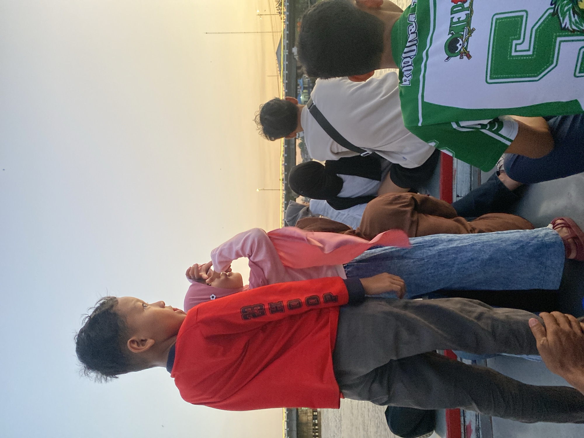 The image captures a group of people, including children and adults, on a boat on the Barito River in Banjarmasin, Indonesia, during sunset. The sky is a soft gradient of pale blue to yellow, indicating the late afternoon. In the foreground, a young boy in a red long-sleeved shirt and grey pants stands facing away from the camera, looking towards the river. Beside him, a girl wearing a pink hijab and light blue jeans also looks away, with her hands near her face. Other individuals are seated on the boat behind them, their backs to the viewer, some wearing casual clothing and one in a green and white jersey with the number '5' visible. A bridge with illuminated lights spans the river in the background, and the water appears calm. The overall atmosphere is one of leisure and observation of the natural surroundings.