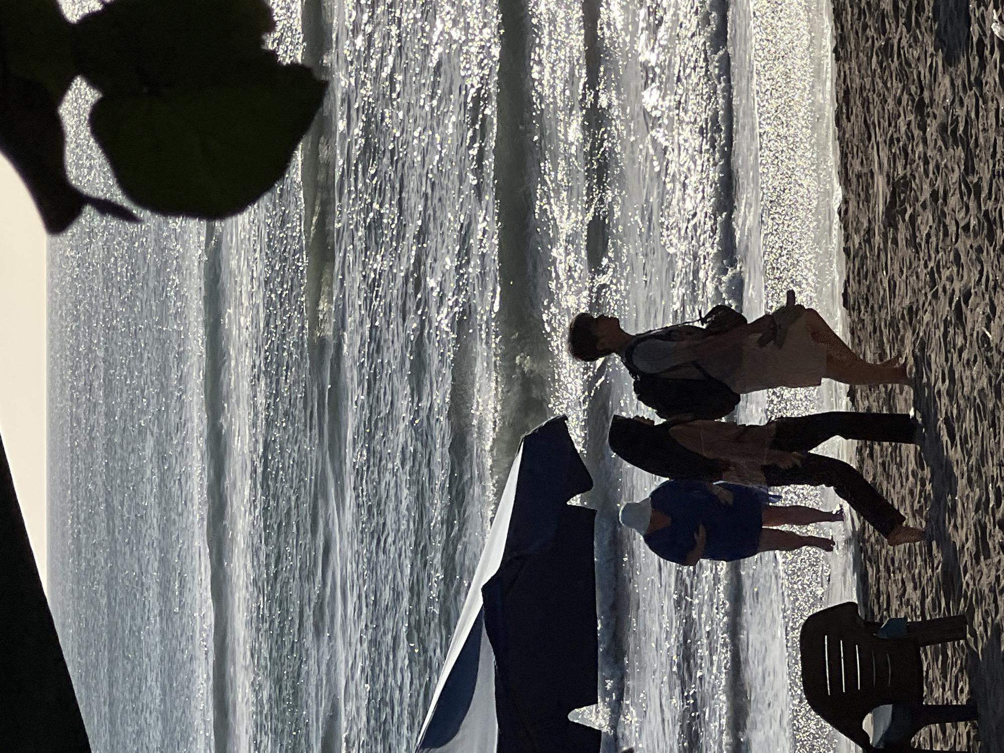 This image depicts a beach scene in Kuta, Indonesia, with the ocean as the primary background. The sun is low, casting a bright, shimmering light on the water, creating a sparkling effect. In the foreground, a sandy beach is visible, with some footprints.
Several people are present:
* On the right, a young person with a backpack is walking along the shore, facing left. They appear to be wearing a light-colored t-shirt and shorts.
* To the left of this person, another individual, likely a woman, is walking towards the right. She has long dark hair and is wearing a light-colored long-sleeved top and dark pants.
* Further to the left, a person in a blue dress and a white hat is walking in the shallow water, heading towards the right.
* A dark blue and white beach umbrella is set up on the sand, partially obscuring the view of the people and the water.
* To the far left, a brown plastic beach chair is positioned on the sand.
* In the upper right corner, a dark silhouette of leaves from a tree is visible, partially framing the scene.
The overall impression is a tranquil beach setting, possibly during the late afternoon or early morning.