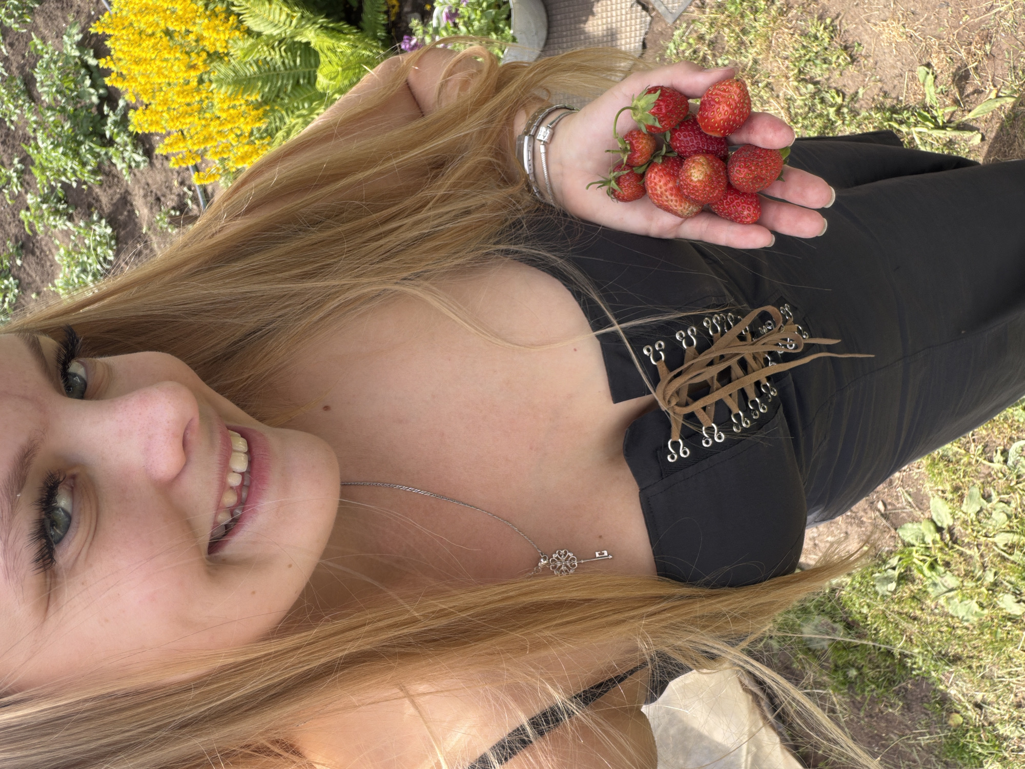 The image features a young woman with long blonde hair, lying outdoors in what appears to be a garden or park setting. She is wearing a black top with a lace-up detail and a silver necklace with a key pendant. Her right hand is holding a small bunch of ripe strawberries. The woman is looking up and smiling, with her face close to the camera, showing her green eyes, freckled skin, and teeth.
In the background, there are green plants, including some with bright yellow flowers and ferns, suggesting a daytime setting during a warm season. The ground is covered with soil and grass. The scene appears to be casual and relaxed, possibly a sunny day, given the bright lighting. No visible text is present in the image.