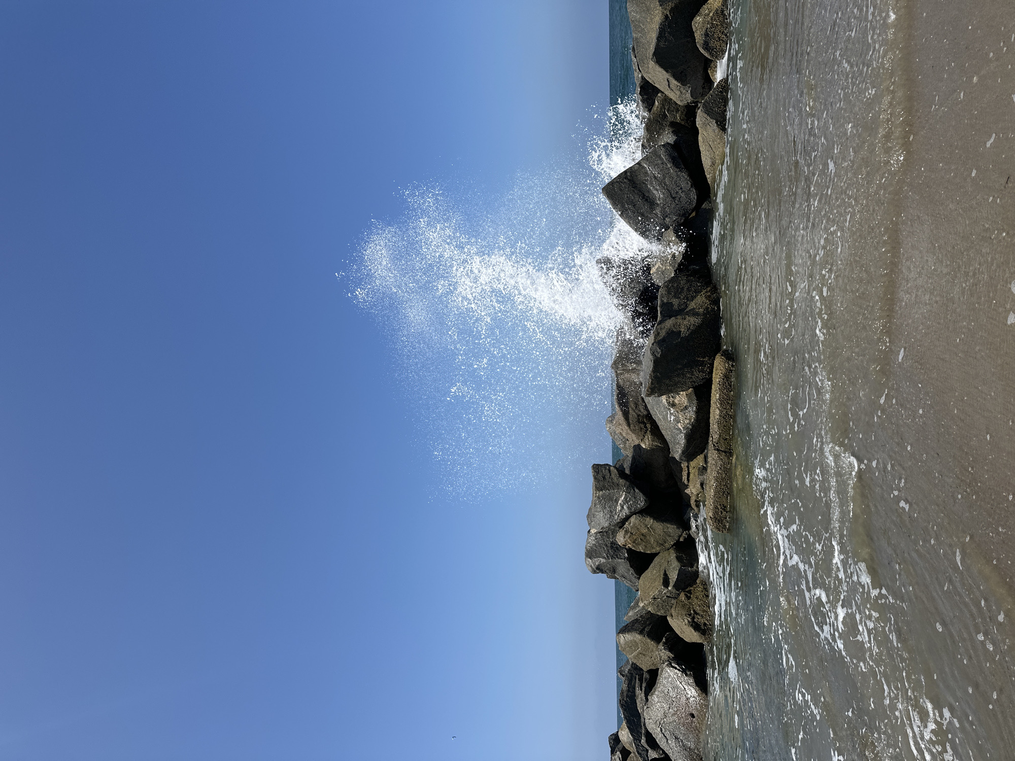 A wave crashes against a rocky breakwater under a clear blue sky. The breakwater is composed of large, dark grey rocks. The water in front of the breakwater is shallow and appears murky, with small waves and foam. The ocean extends to the horizon in the background, where it meets the sky. The scene appears to be a coastal area, possibly a beach or a harbor entrance. The image is taken from a low angle, looking slightly up at the breakwater and the crashing wave. There are no people or animals visible in the image. The overall impression is one of natural power and the dynamic interaction between water and land.