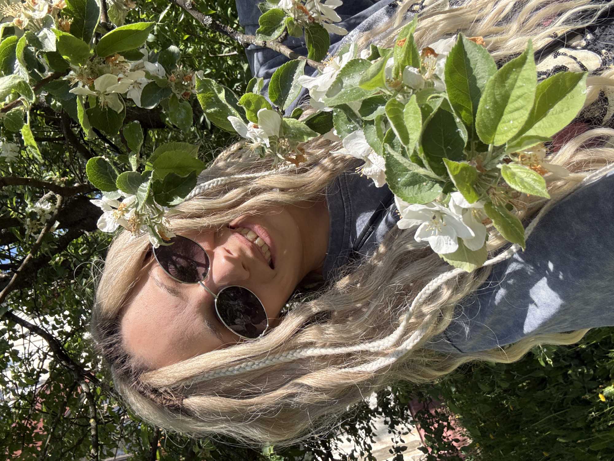 A woman with blond dreadlocks and round sunglasses is smiling while positioned under a tree with white flowers and green leaves. She is wearing a denim jacket. The background is filled with foliage and glimpses of the sky. The location is Rusanovo, Russia.