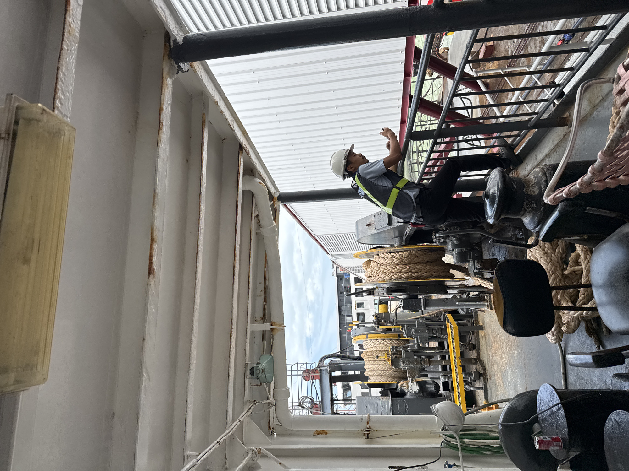 The image depicts a worker on the deck of a ferry or ship, likely in a port or dock area. The worker, wearing a white hard hat and a high-visibility vest over a gray shirt and dark pants, is leaning on a railing and looking at a mobile phone. In the background, large ropes are coiled around winches, suggesting equipment for mooring or cargo handling. Various pipes and metal structures are visible, indicating an industrial or maritime setting. The overall environment appears somewhat weathered, with some rust spots visible on the overhead beams and metalwork. The scene suggests a moment of downtime or a check-in during a work shift on a vessel. There are no specific indicators to pinpoint the location to a particular city in Thailand; it's a general maritime scene.