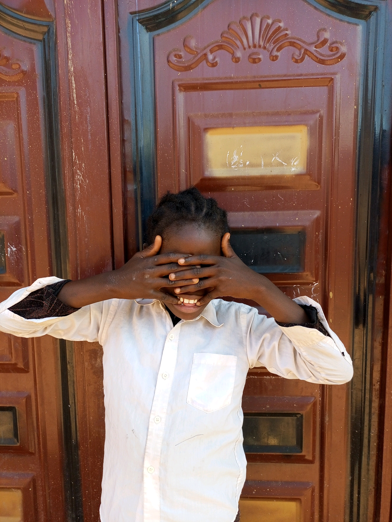 A young person is standing in front of a brown double door with decorative panels and a narrow glass window in the upper panel. The person is wearing a light-colored collared shirt and has their hands covering their eyes, with their mouth open in a smile. Their arms are outstretched. The door appears weathered and has some white speckling on its surface. The image is taken outdoors.