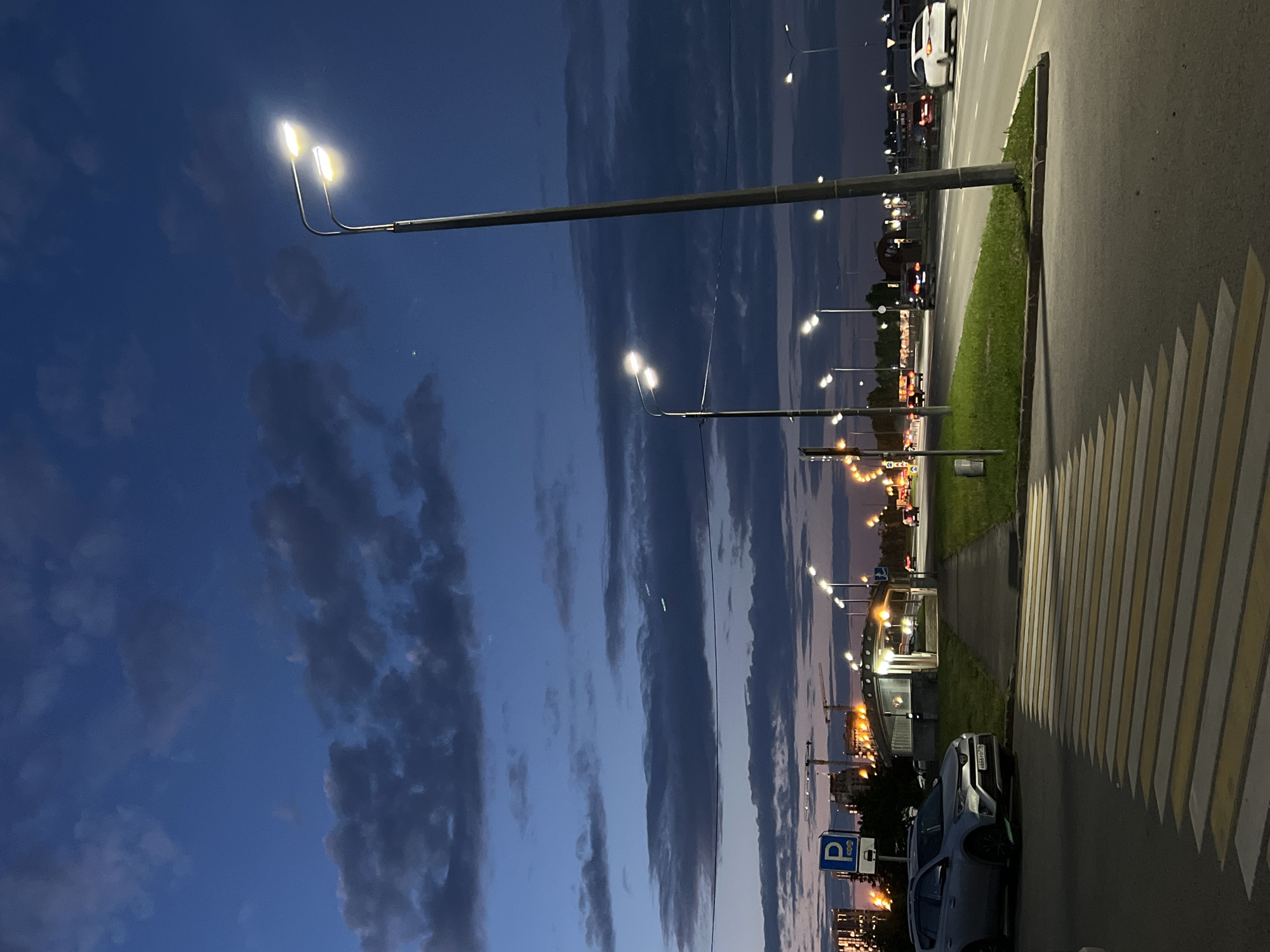 The image captures a twilight scene in Kazan, Russia, likely near a residential or commercial area, indicated by apartment buildings and construction cranes visible in the distance. A silver SUV is parked to the left, near a pedestrian crosswalk. A sidewalk leads to a small, well-lit pavilion. Streetlights illuminate the road, where several cars are driving. The sky is a deep blue with scattered dark clouds, suggesting the transition from day to night.