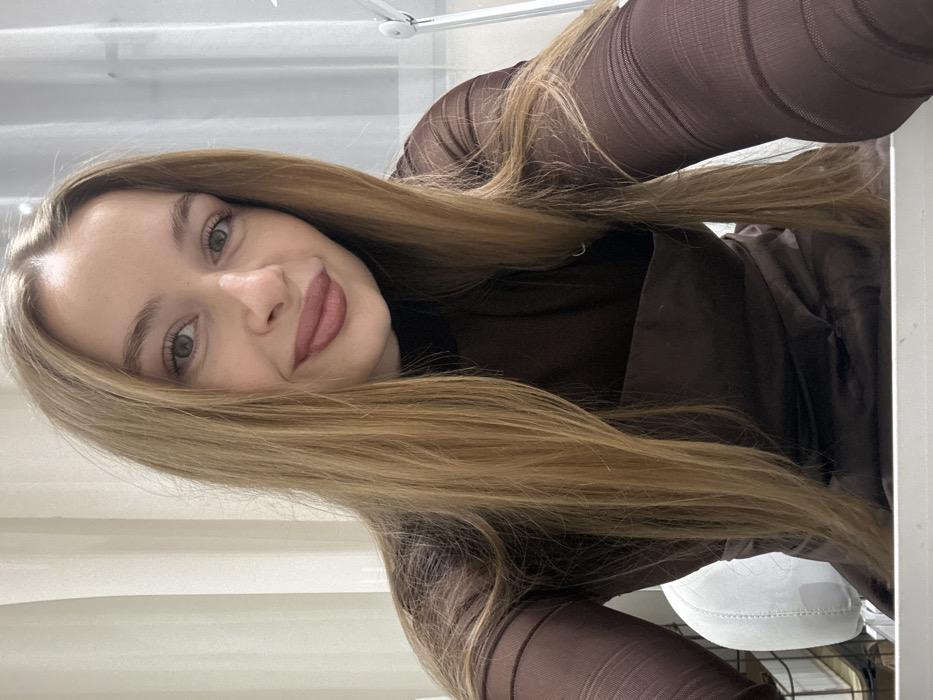 A young adult female is depicted from the upper torso upwards in an indoor setting. She has long, straight, light brown hair parted in the middle, extending over her shoulders. Her eyes are light green/hazel, and she has a slight smile with lips colored by a matte, plum-toned lipstick. She is wearing a dark brown, possibly sheer or mesh, long-sleeved top, layered over a darker, opaque dark brown garment underneath, which appears to be a dress or camisole with a straight neckline.
The background shows an interior room, well-lit with artificial lighting. To the subject's left, a section of a white or off-white curtain is visible, along with the top portion of a white, possibly upholstered, chair back. To the subject's right, a window with a sheer white curtain or blind is partially visible, alongside a silver or white floor lamp with an adjustable arm. A white, flat surface, likely a desk or table, is present at the very bottom edge of the frame. The specific location within Budva, Montenegro, is not discernible from the visual evidence, but the scene is an indoor room. The subject is looking directly at the camera.