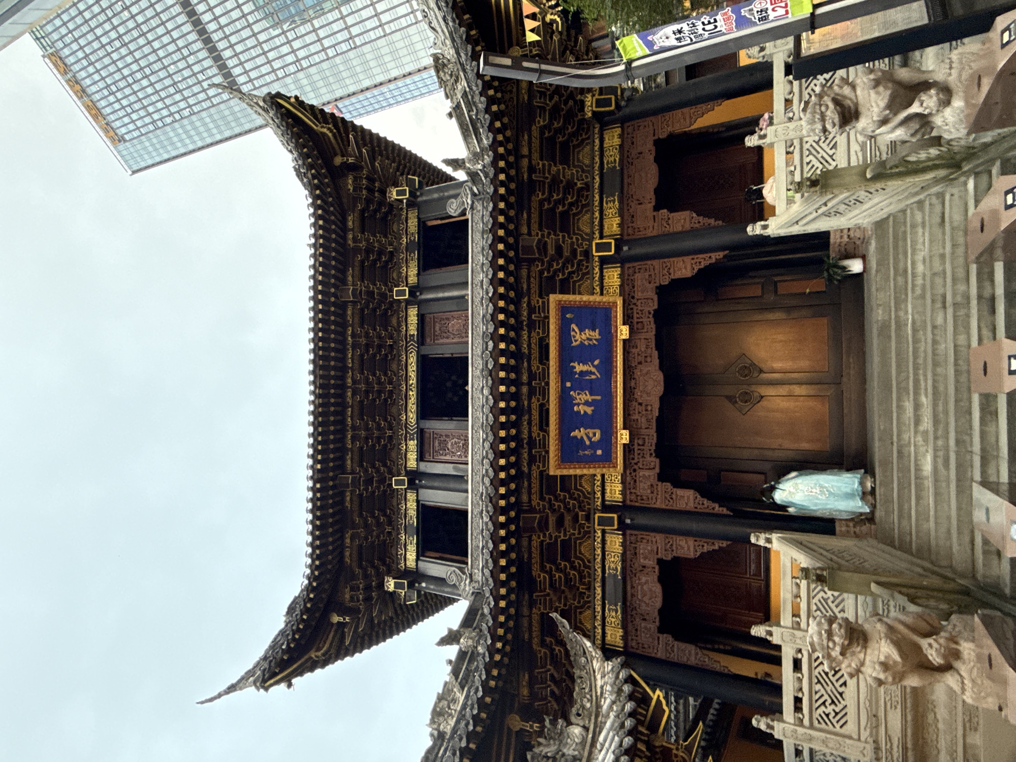 An exterior shot captures the ornate entrance of a traditional Chinese temple, possibly located in Yuzhong District, China. The temple features a multi-tiered, dark wooden roof with intricate golden detailing and upturned eaves. A blue plaque with gold Chinese characters hangs above a large, closed wooden door. Stone lion statues flank the wide stone staircase leading up to the entrance. A person in a flowing light blue garment stands on the stairs, facing away from the camera and towards the temple entrance. In the background, a modern glass skyscraper rises high into the cloudy sky, contrasting with the ancient architecture of the temple. A vertical banner with Chinese text and imagery is attached to a pole on the right side of the frame.