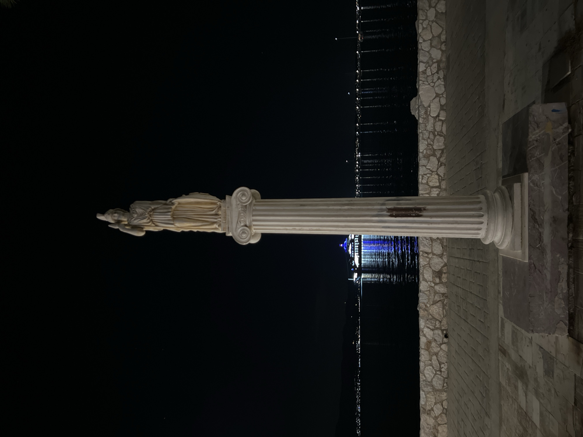 A nighttime, medium shot captures a classical-style column topped with a statue, positioned on a paved walkway in Eretria, Greece. The statue depicts a draped female figure, possibly Athena, holding an object in her right hand. The column itself is fluted and made of a light-colored material, possibly marble. In the background, a body of water reflects the lights of a city and a large ship illuminated with blue and white lights. The water is calm, with a stone wall lining the walkway. The sky is dark and clear, with a few visible stars. The scene is likely a public promenade or waterfront area.