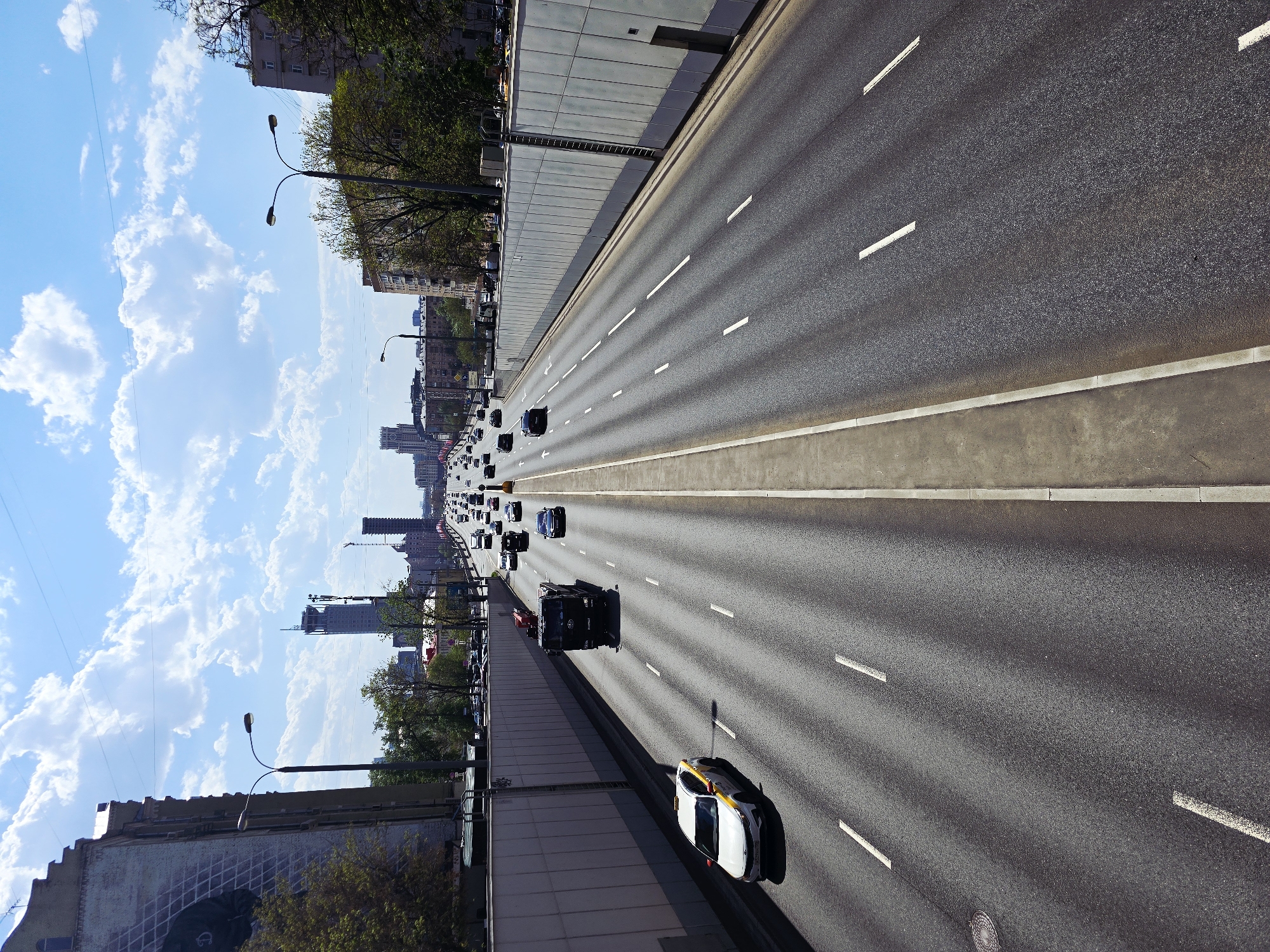 The image depicts a wide, multi-lane highway in Moscow, Russia. The view is from an elevated position, looking down the highway with a slight perspective. Cars are traveling in both directions, with a taxi cab prominently featured in the foreground. Buildings and trees line the side of the highway, contrasting with the bright blue sky filled with puffy clouds. Streetlights are visible along the roadway. The highway is marked with white lines indicating lanes.