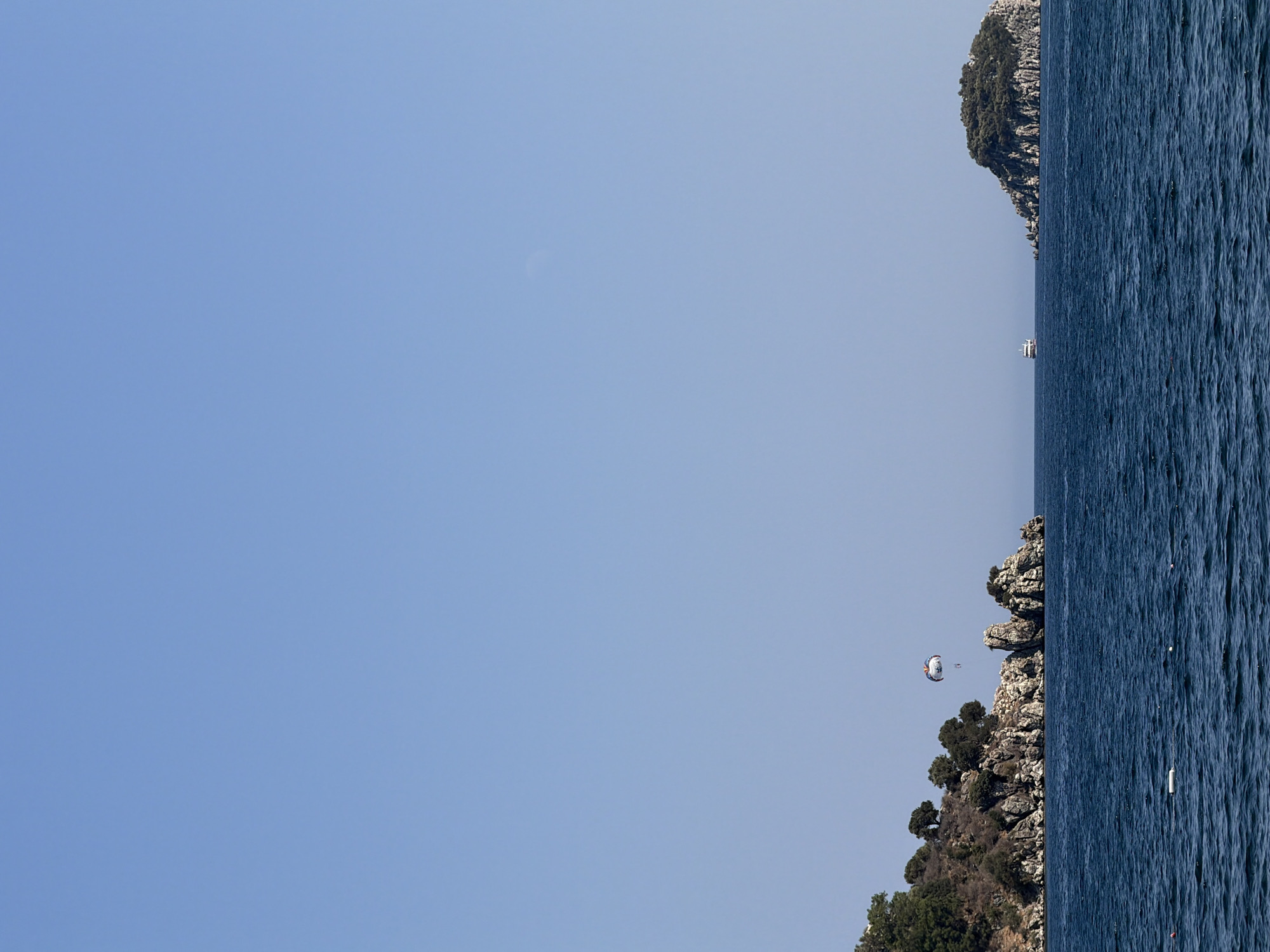 This image captures a scenic view of the Aegean Sea near Marmaris, Turkey, under a clear blue sky. The foreground is dominated by deep blue, rippling water, with a few buoys visible on the surface.
In the middle ground, a rocky, tree-covered island juts out from the sea on the left side of the frame. On this island, a parasail with a colorful canopy is visible in the air, with two figures beneath it, indicative of a recreational activity. Nearby, large rock formations break the waterline. Further to the right, a white boat is seen sailing on the sea.
On the right side of the image, a smaller, similarly rocky and tree-covered island is situated, appearing as a distant landmass. The overall scene conveys a sense of a tranquil yet active coastal environment, characteristic of popular tourist destinations like Marmaris.