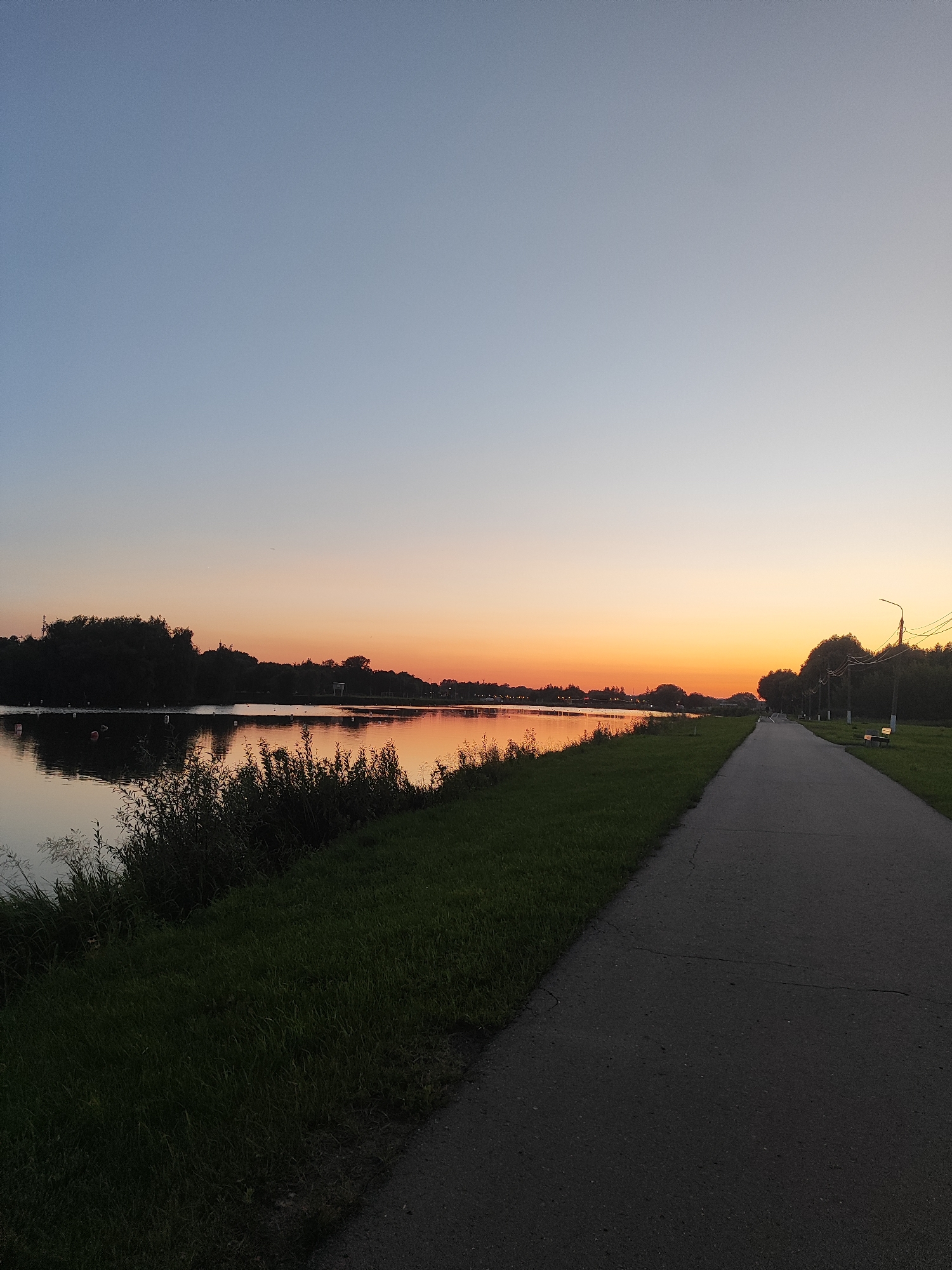 The image depicts a sunset over a lake in Bronnitsy, Russia. A paved pathway runs along the edge of the lake, bordered by a grassy area. Trees line the opposite side of the lake, their silhouettes dark against the colorful sky. The water reflects the hues of the sunset, creating a serene and picturesque scene. A single bench is visible on the pathway, suggesting a public park or recreational area. There are no people or animals visible in the frame. The overall atmosphere is tranquil and peaceful, characteristic of a quiet evening in a natural setting.