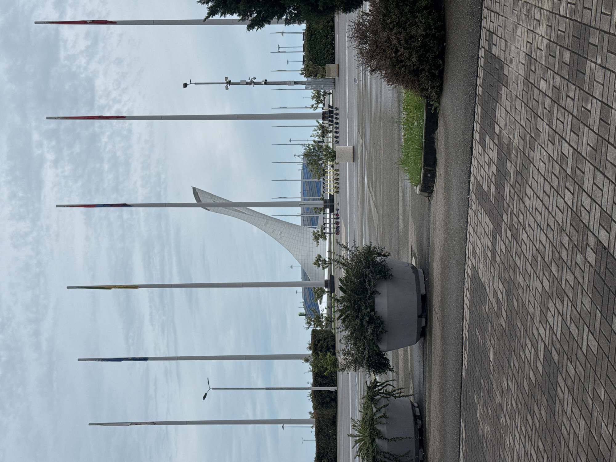The image depicts an outdoor urban scene in Sirius, Russia. A paved area, possibly a sidewalk, is visible on the right with a gray brick pattern. Adjacent to the paved area is a grey, wet road or walkway lined with concrete planters containing bushes and greenery.
Along the left side of the scene are multiple tall, white flagpoles. Most of them have a remnant of colored fabric at the top, suggesting they previously held flags. In the background, there is a large, white, arched architectural structure. In the far background, a low building with a blue roof can be seen.
The sky is overcast with a layer of clouds. There are also streetlight poles of varying sizes visible. The image is oriented vertically. There do not appear to be any people in the scene.