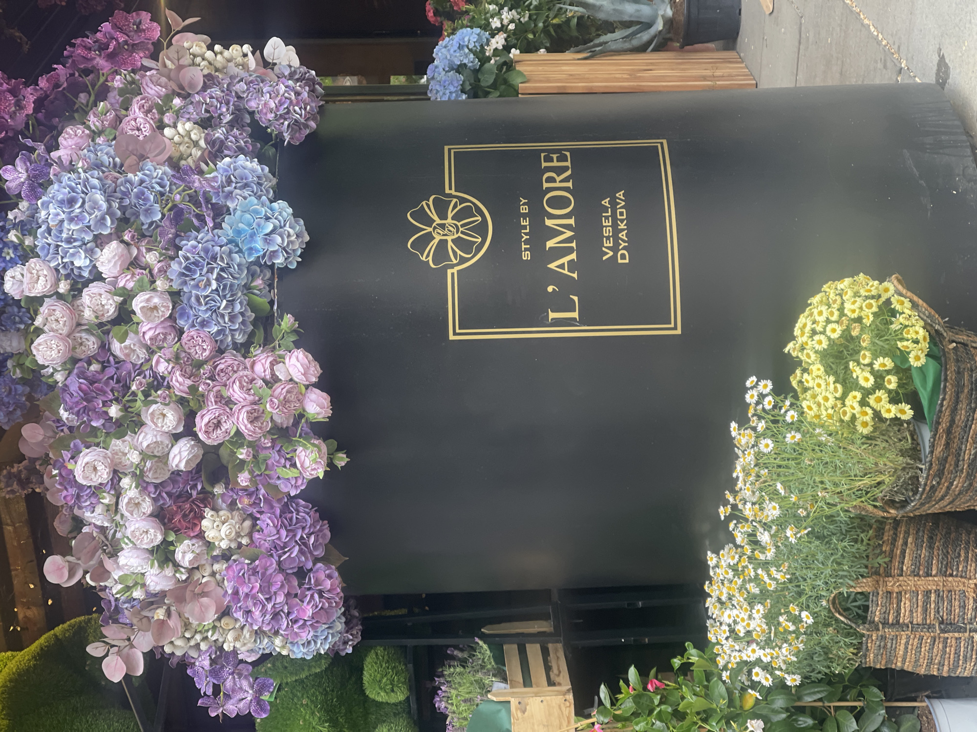 A flower arrangement advertisement in Sofia, Bulgaria, features a black cylindrical backdrop with a gold-colored framed logo on its front side that reads "STYLE BY L'AMORE VESELA DYAKOVA." Artificial floral arrangements in shades of purple, pink, blue, and white are positioned on the left side of the cylinder. On the right side are flower arrangements of white and yellow daisy-like flowers in woven baskets. A variety of other greenery is seen below the cylinder. The sidewalk is visible at the top right corner of the image.