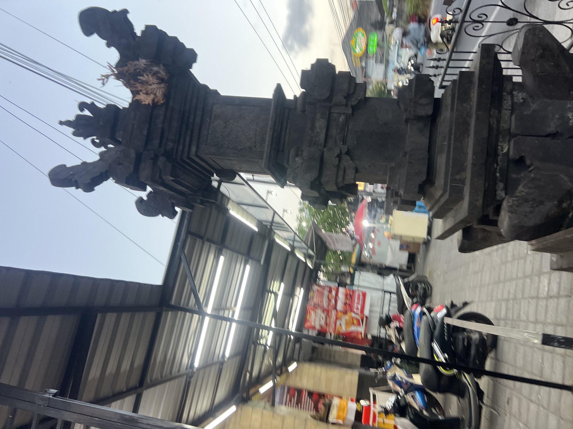 The image shows an outdoor scene in Denpasar, Indonesia, likely in a commercial area. In the foreground, a dark stone statue with intricate carvings stands prominently. Behind the statue, a covered walkway with metal roofing and fluorescent lighting lines the side of a building. Several motorcycles are parked along the walkway. Further in the background, street vendors with food stalls and signage are visible. A road with traffic, including cars and motorcycles, runs alongside the walkway. The sky is overcast, suggesting it is either early morning or late afternoon. The overall impression is one of a bustling marketplace or street with a blend of traditional and modern elements.