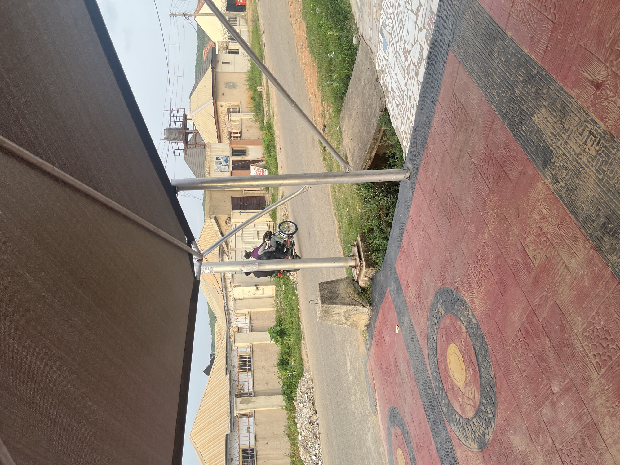 In Abuja, Nigeria, the image captures a street scene from under a large, light brown awning. A paved walkway with a red and black patterned surface is in the foreground. Beyond the walkway, a two-lane asphalt road stretches into the distance. On the road, two individuals are riding a motorcycle, with the rider wearing a dark shirt and the passenger a purple shirt.
To the left of the road, there is a pile of construction debris and a patch of overgrown green grass. A concrete block with a U-shaped metal bracket is positioned near the edge of the road.
The background features several single-story buildings with beige walls and corrugated metal roofs. Some of the buildings have security bars on their windows and barbed wire along their fences. A water tower with a metal platform is visible in the distance behind one of the buildings, along with power lines and utility poles. The sky is overcast. The overall impression is of a residential or commercial area in an urban setting.