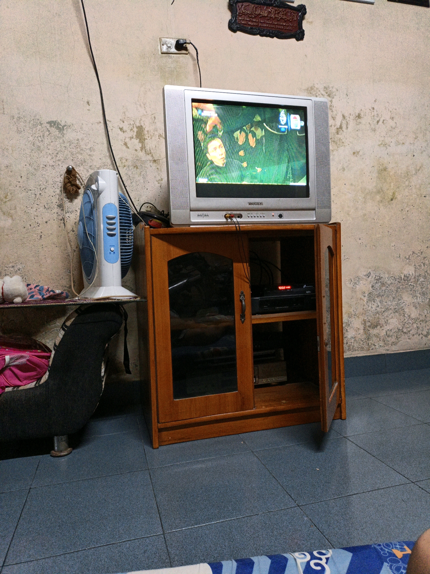In the city of Bunut, Indonesia, a vintage Toshiba television set displays a scene from a television program. The television rests on a wooden cabinet with an open door, revealing a black electronic device with a red digital display reading "1002." To the left of the television, a blue and white oscillating fan stands on a shelf alongside a stuffed toy. In the background, a beige wall is adorned with a black framed Islamic calligraphy plaque and a power outlet with a black cord plugged into it. The floor is tiled in a dark blue color. A portion of a dark-colored sofa with visible metal legs is visible to the left of the cabinet. A person's leg is visible in the bottom right corner of the image.