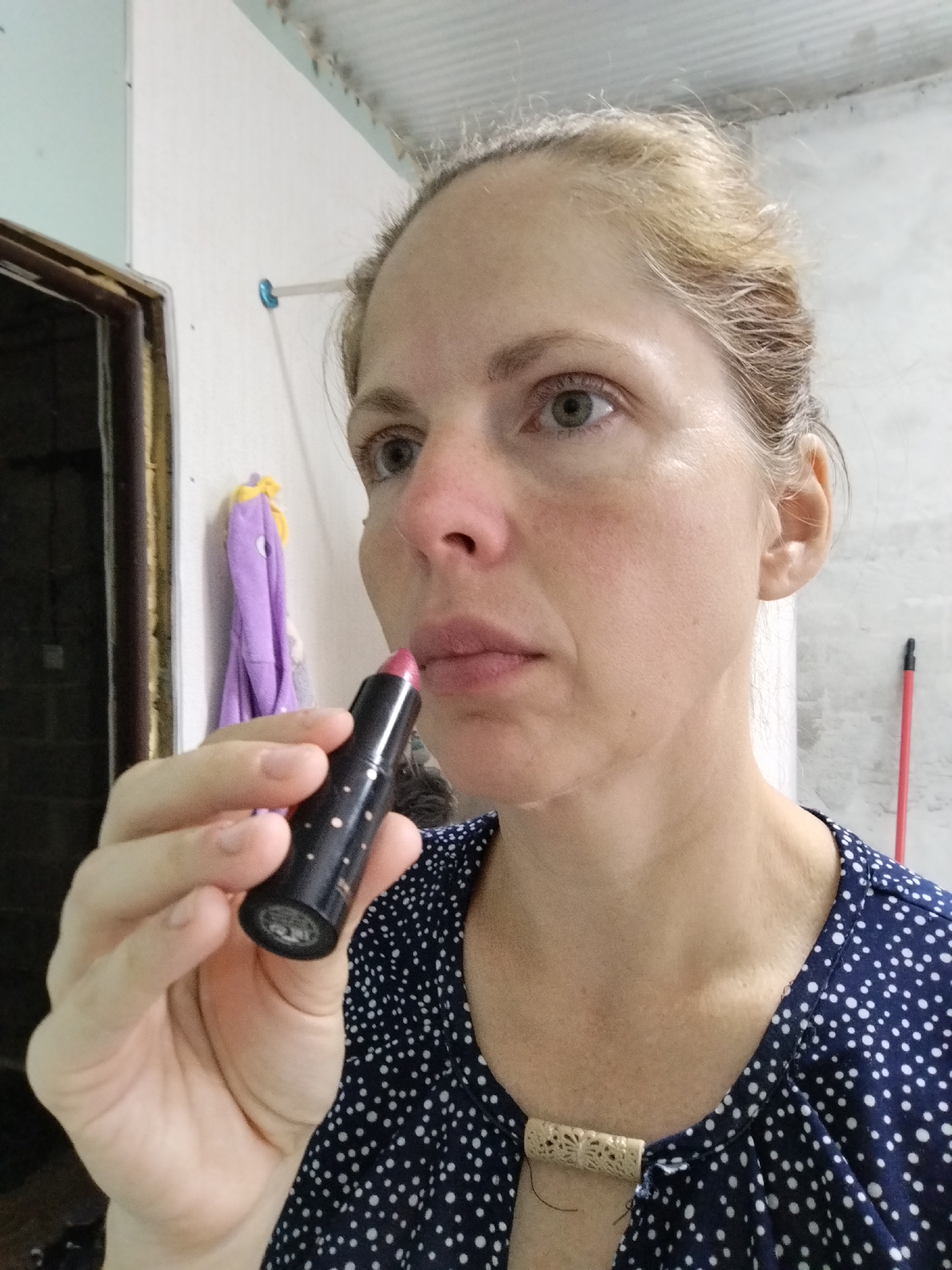 A medium shot shows a Caucasian woman with blonde hair pulled back, applying pink lipstick from a black tube with white polka dots. She is wearing a navy blue dress with white polka dots and a decorative, cream-colored clasp at the neckline. Her left hand holds the lipstick, positioned near her mouth. The background includes a white wall, a partially open dark doorway, a purple cloth hanging on the wall, and a red-handled broom leaning against the wall on the right. The ceiling appears to be corrugated metal. The scene suggests an indoor setting, possibly a bathroom or bedroom, with unfinished or rustic elements.
