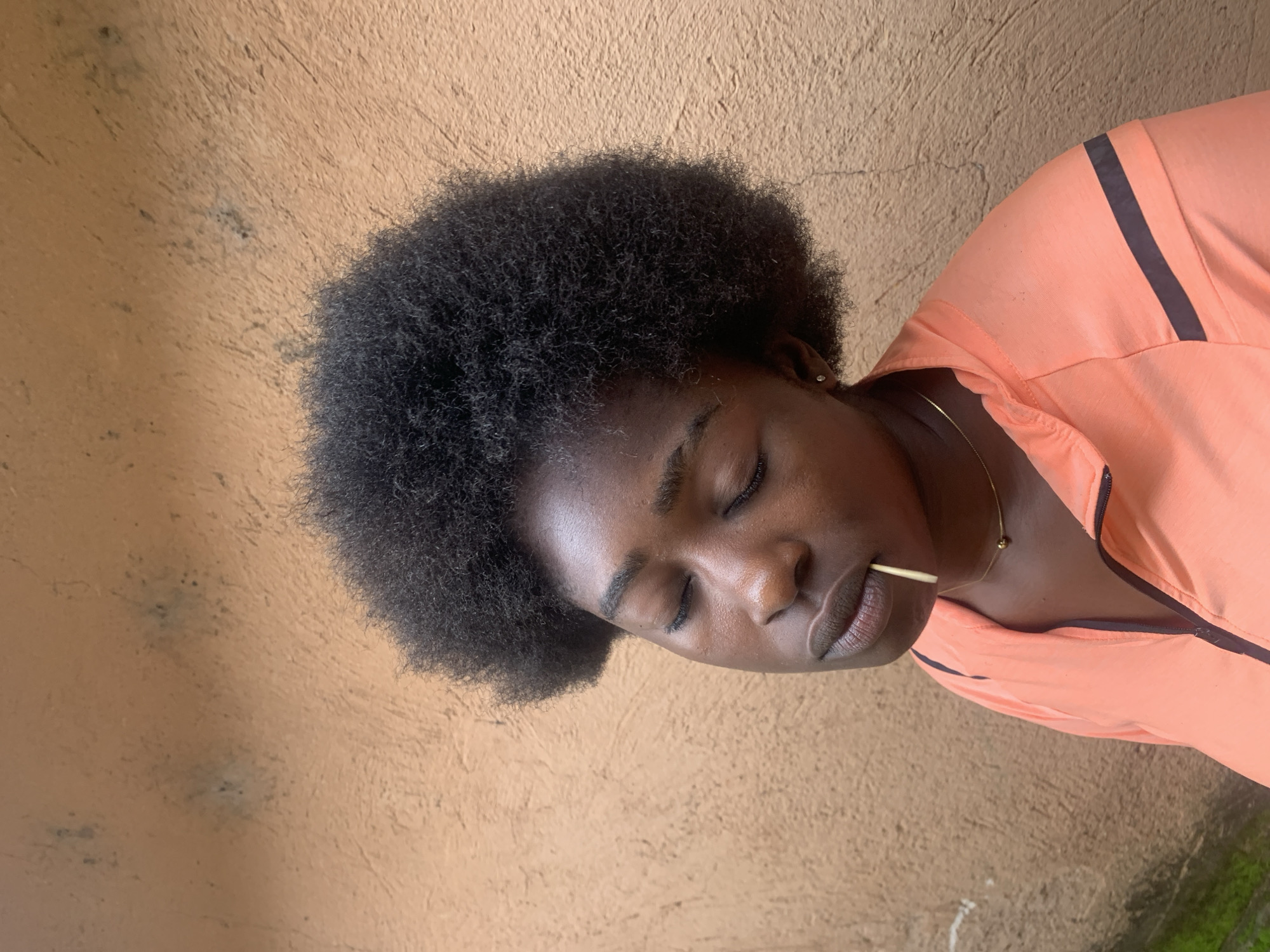 A young Black woman with dark, voluminous natural hair is shown from the chest up, standing in front of a textured, light brown wall. Her eyes are closed, and she has a neutral expression. A thin, light-colored object, possibly a toothpick or a straw, is held between her lips. She is wearing an orange zip-up jacket with dark stripes on the shoulders. A delicate gold chain with a small dark bead is visible around her neck. The background wall shows signs of weathering and some dark spots. The lighting suggests an outdoor setting during the day. The location is identified as Rafin Par, Nigeria.