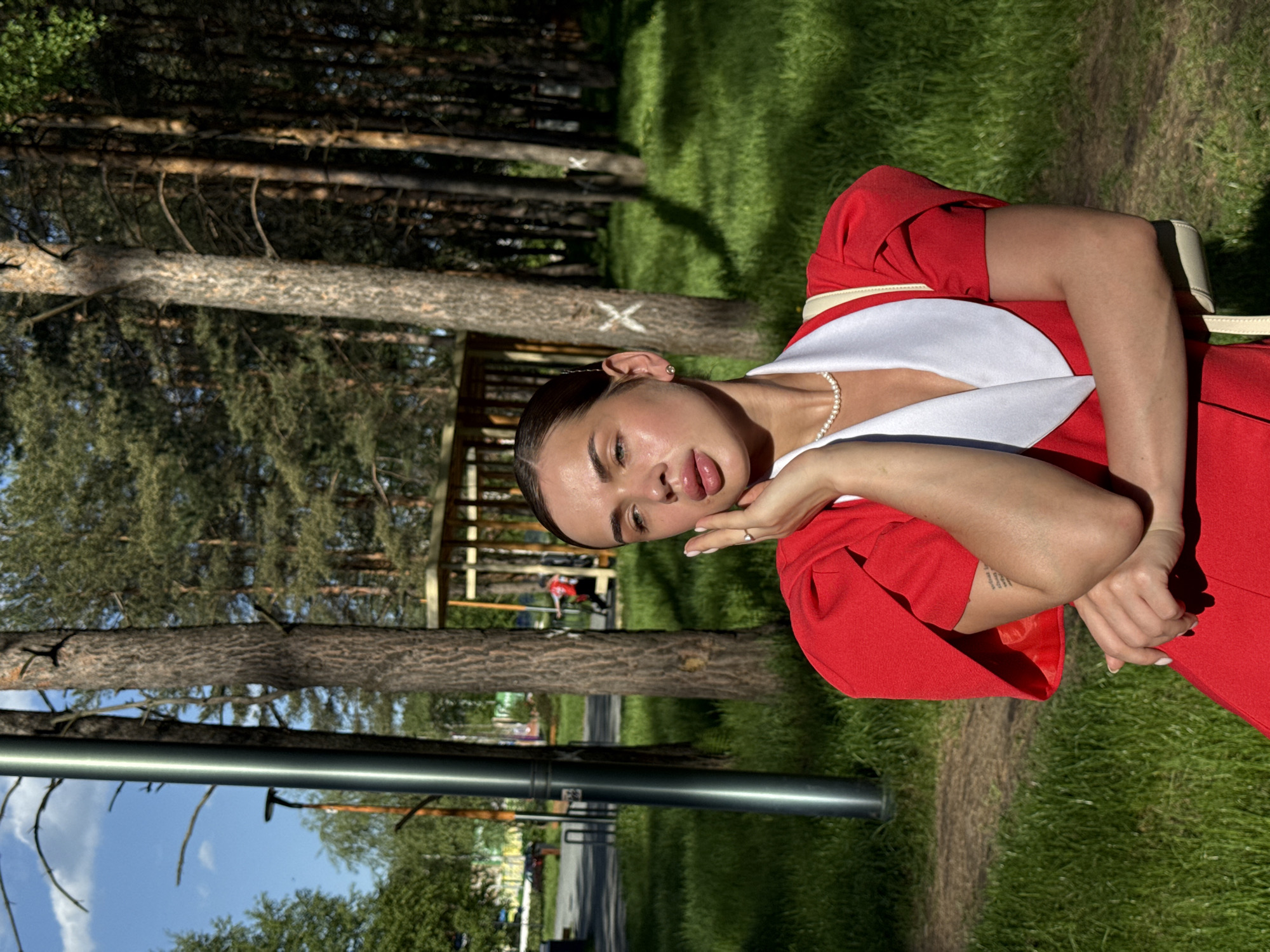 A woman in a red and white dress poses in a park in Perm, Russia. The location has trees, grass, and walking paths.