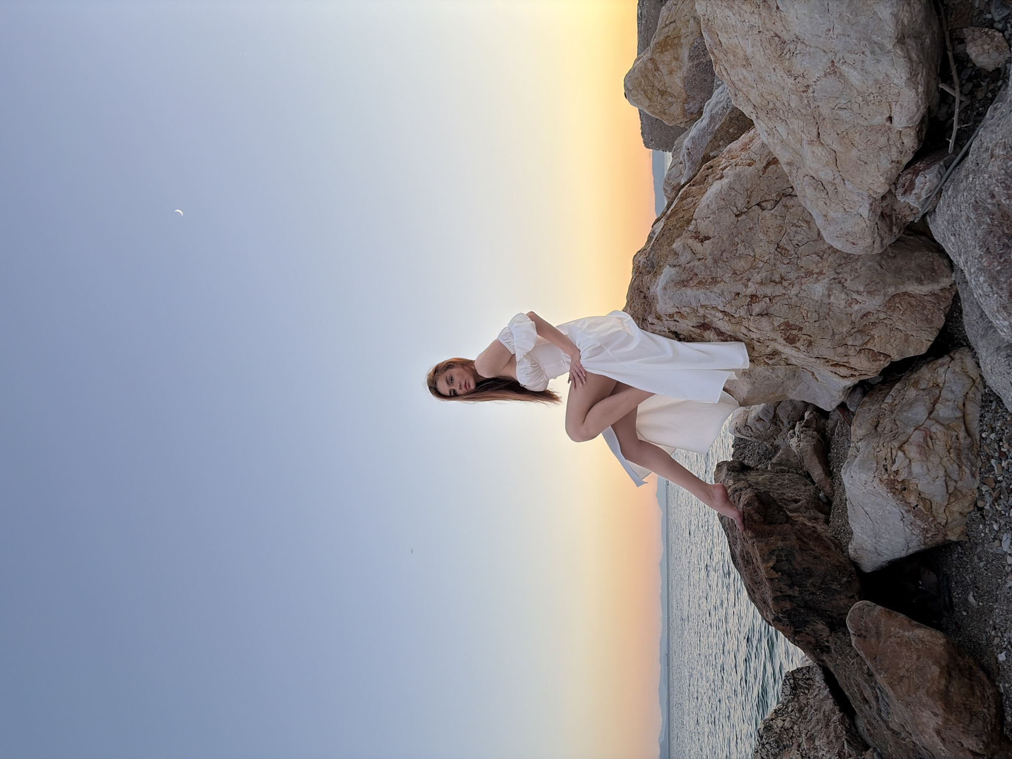 A young woman with long, brown hair is seated on large, light-colored rocks near a body of water. She is wearing a white, off-the-shoulder dress with puffed sleeves and a high slit. The woman is looking towards the camera. The water is calm and reflects the orange and yellow hues of the sunset. The sky is a gradient of blue and orange, with a crescent moon visible in the upper right quadrant. Mountains are visible in the distance across the water. The rocks appear to be part of a breakwater or coastal path, consistent with the coastline of Paleo Faliro, Greece. The overall scene is tranquil and bathed in the warm light of dusk.