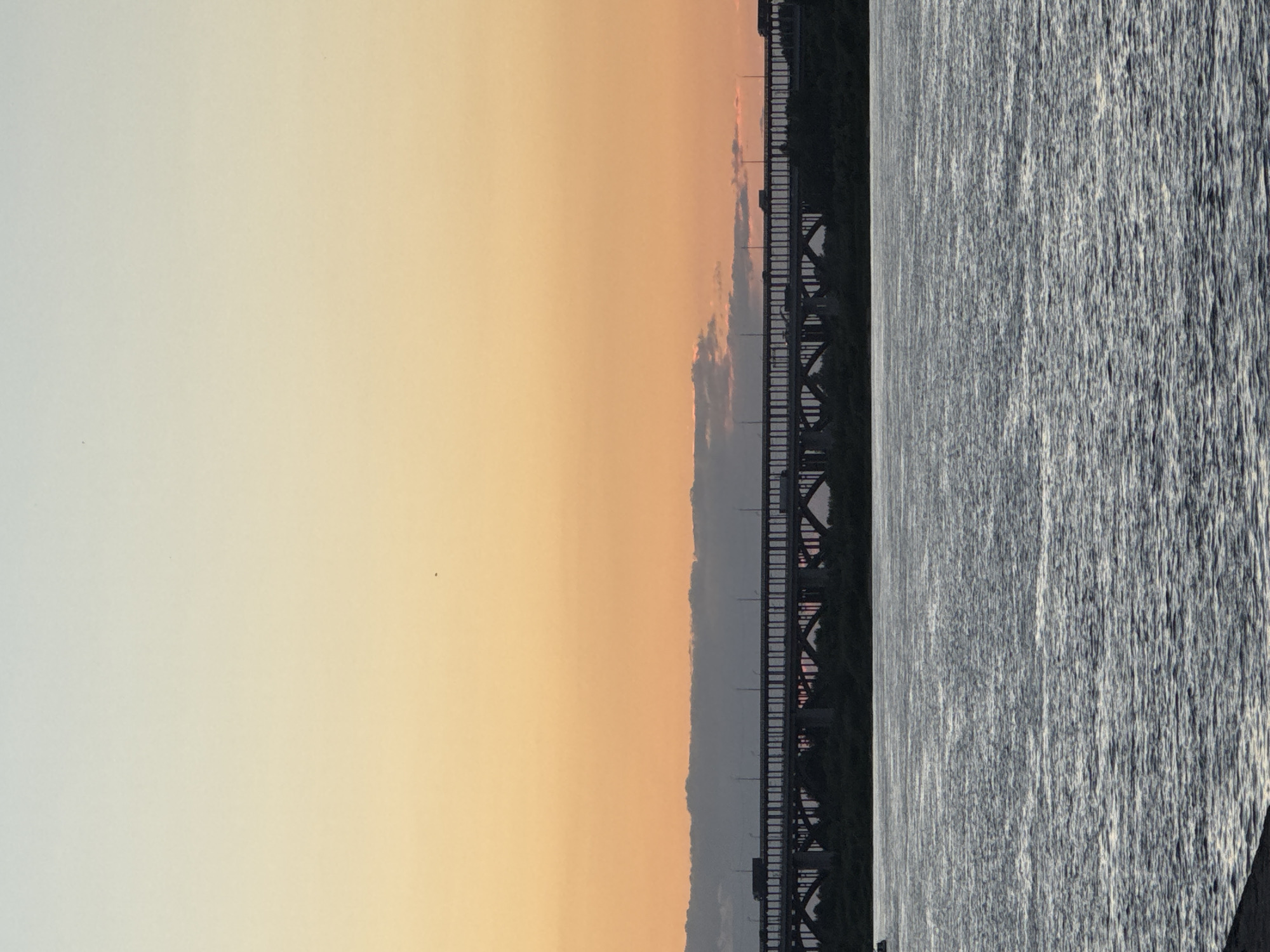 The image depicts a bridge spanning across a body of water, likely the Volga River, in Nizhny Novgorod, Russia. The scene is captured during sunset, with the sky transitioning from a soft orange hue near the horizon to a pale yellow and then a light blue further up. A prominent metal truss bridge stretches across the frame, its dark silhouette contrasting with the warm sky. The water below the bridge appears textured and rippling, reflecting the muted colors of the sky and the industrial structure. The overall mood is serene and peaceful, evoking the tranquility of a day's end. There is no visible text in the image.