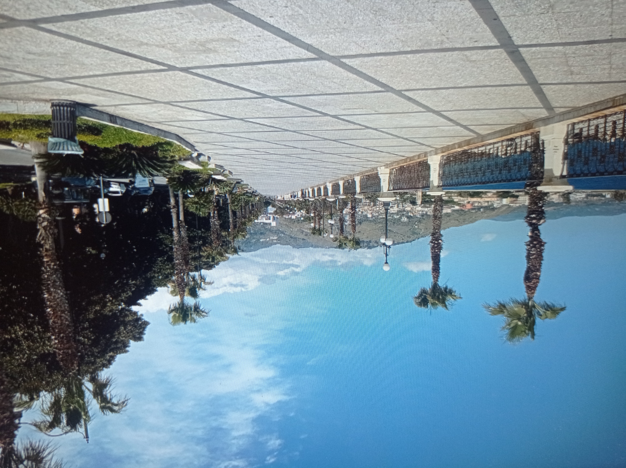 The image depicts a wide, paved promenade lined with palm trees and a railing overlooking a body of water, likely the sea or a large lake. The promenade is paved with large, square tiles, casting geometric shadows. In the background, there are hills with buildings scattered across them, suggesting a coastal or lakeside town. The sky is clear blue with a few wispy clouds. There are no people clearly visible in the foreground, but there are some figures in the distance walking along the promenade. The scene suggests a pleasant, sunny day in a tourist area. This image is not from Mukachevo, Ukraine; it appears to be a Mediterranean coastal location.