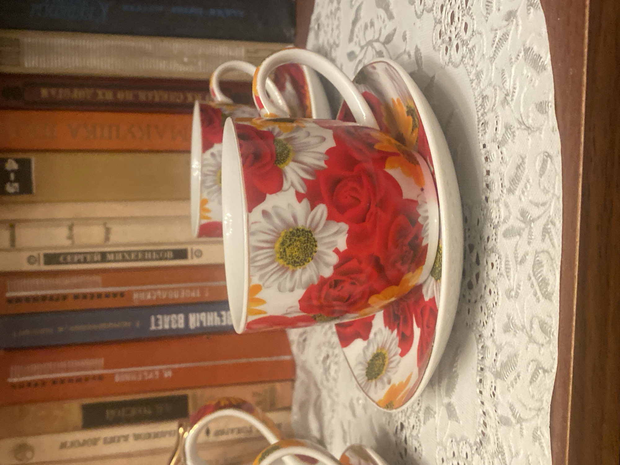 The image displays a close-up view of two floral-patterned teacups and saucers, positioned on a white lace doily. The teacups are adorned with red roses and white daisies. In the background, a bookshelf filled with books is visible. The lighting suggests an indoor setting. The location of Novaya, Russia is not discernible from the visual information presented.