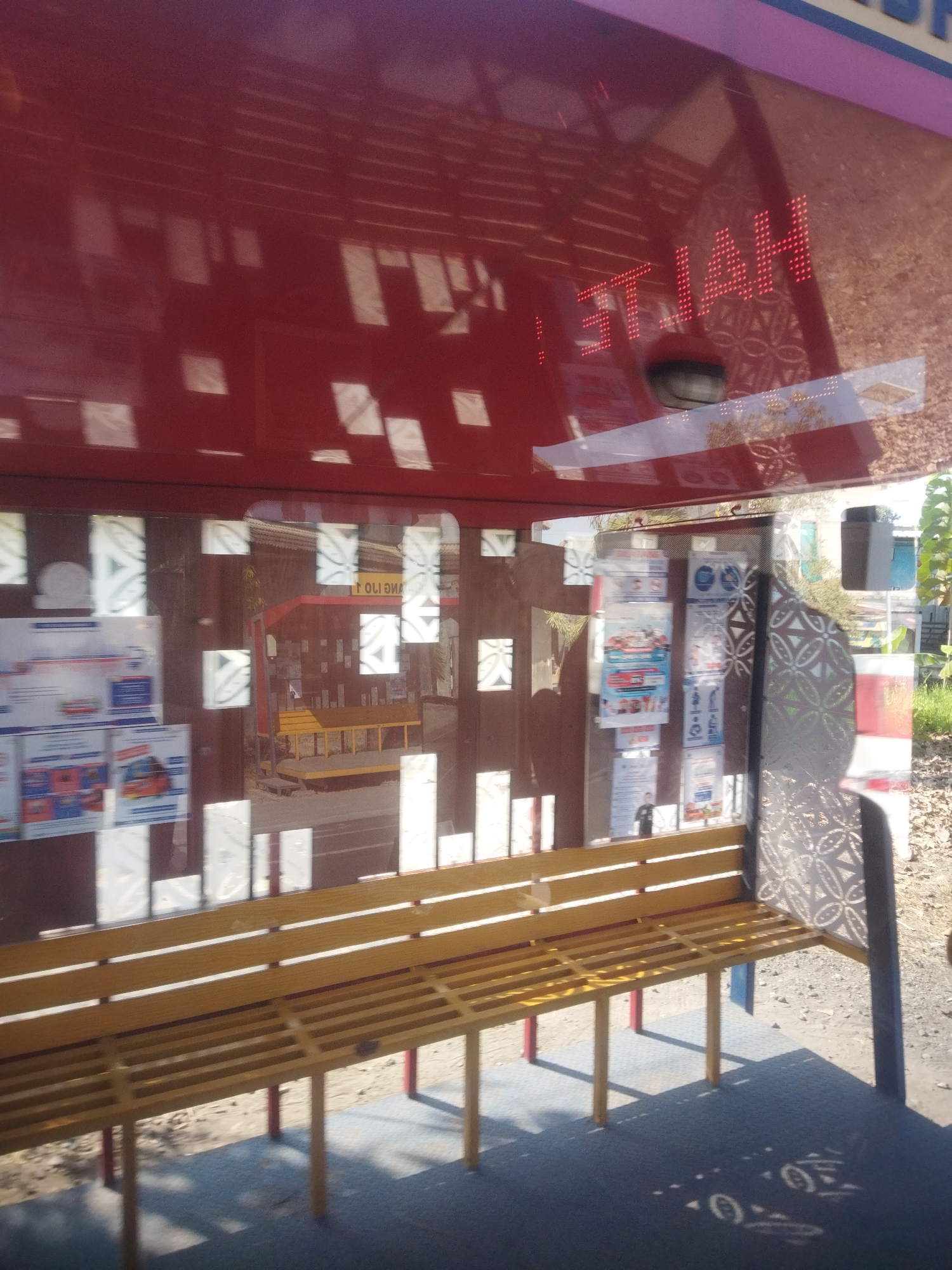 The image shows a bus stop shelter in Nambangan, Indonesia. The shelter has a red roof and sides, with glass panels that reflect the surroundings. Inside the shelter, there is a yellow bench with red legs. A red sign with white lettering is visible in the background, which appears to be some form of advertisement or announcement. The ground is paved with blue tiles and gravel. The lighting suggests it is daytime with sunlight casting shadows.
