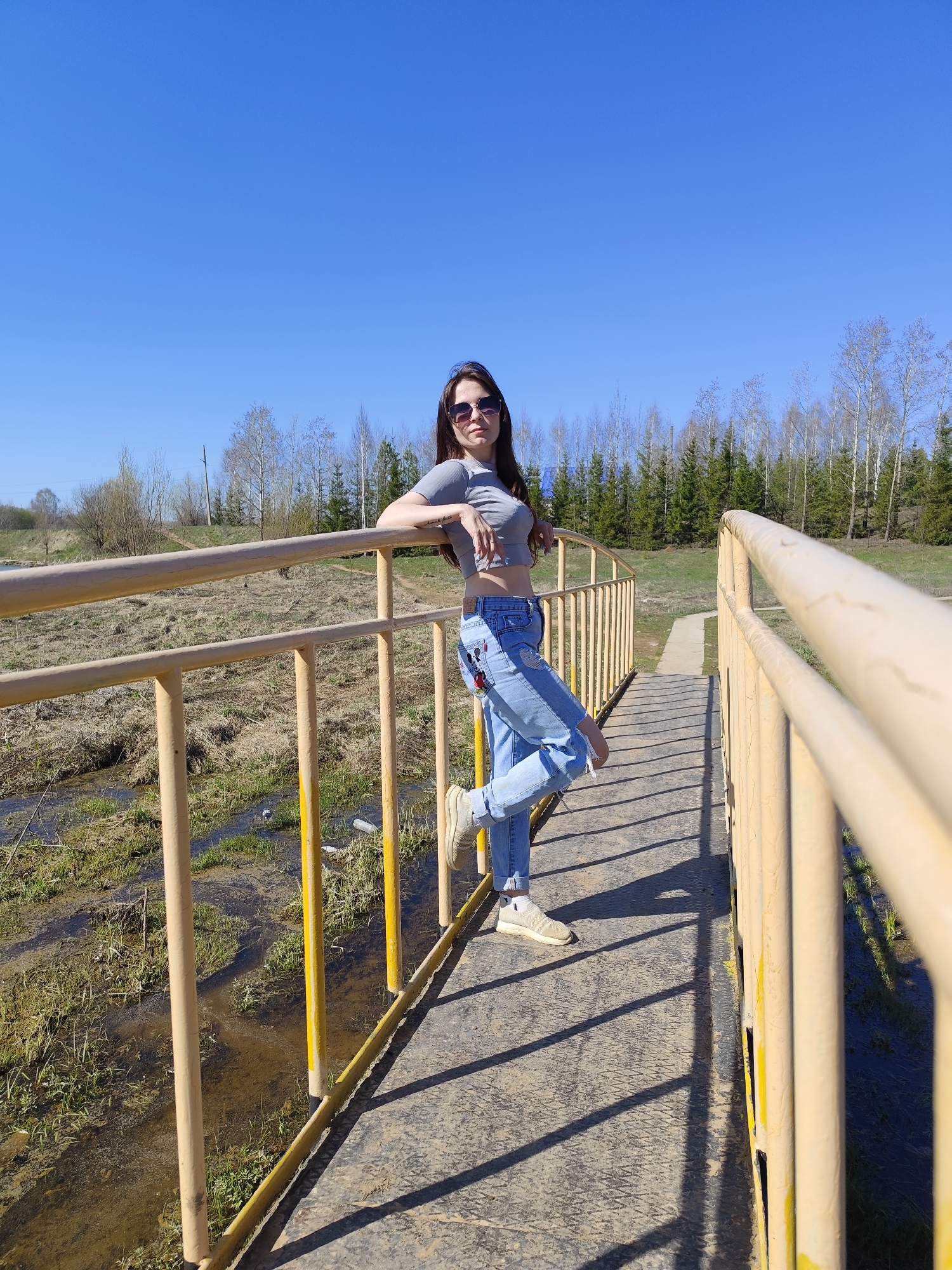 A young woman is standing on a yellow bridge in Фоки, Russia. She is wearing a gray crop top, light blue jeans, and sneakers, and she has sunglasses on. She is leaning against the railing of the bridge. The bridge crosses a grassy area with water flowing underneath. In the background, there is a forest of birch and evergreen trees under a clear blue sky.