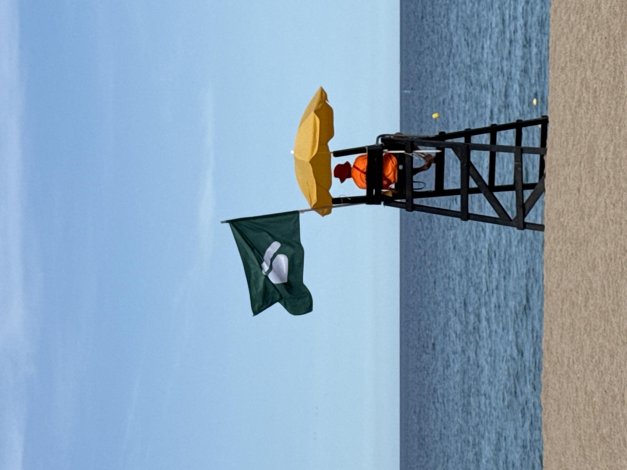 The media file shows a lifeguard stand on a beach, with the ocean and sand visible. A person in an orange shirt and red hat, likely a lifeguard, is seated on the stand, under a yellow umbrella. A green flag with a white symbol is attached to the stand. The scene appears to be set during the day, with clear blue skies and calm water. The image is oriented vertically, suggesting it was taken with a phone camera. The presence of a lifeguard and the beach setting indicates a recreational area, likely during warmer weather. No visible text is present in the image. The location context provided is Blanes, Spain, which is a coastal town known for its beaches.