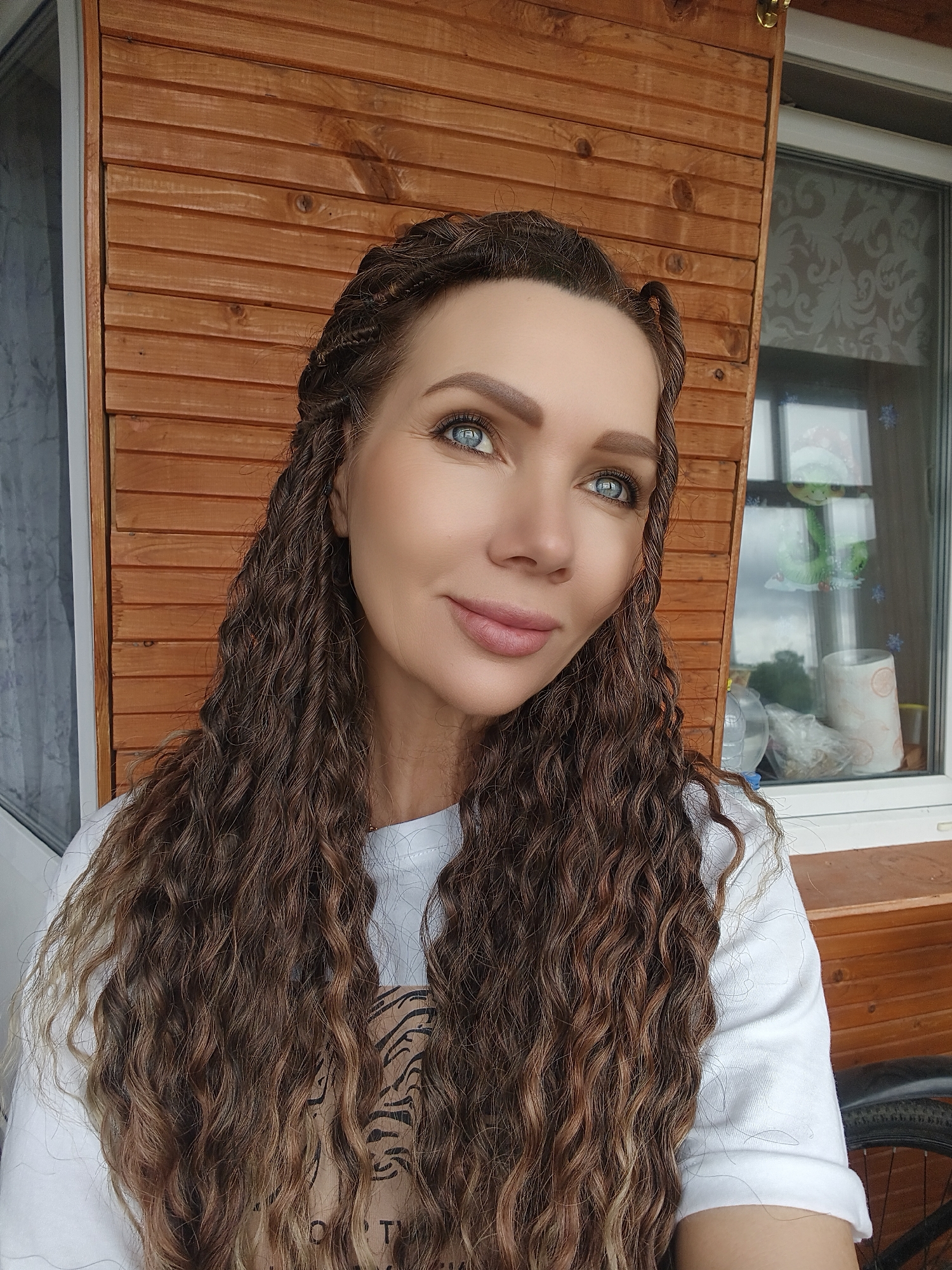 A close-up, mid-shot portrait features an adult female situated in the city of Unknown, Unknown. She possesses prominent bright blue eyes, defined eyebrows, and light pink lipstick on full lips, exhibiting a slight upward gaze and subtle smile. Her long, wavy-curly brown hair, featuring lighter highlights, extends past her shoulders, with sections around her face styled into tight twists or braids. She is wearing a white short-sleeved t-shirt with a visible dark graphic pattern (resembling tiger stripes) on the chest area. The background primarily consists of horizontal wooden plank siding, indicating an outdoor structure such as a porch or cabin wall. To her right, a white-framed window reveals a partially drawn patterned blind or curtain and a view of an overcast sky with some distant greenery. Adorning the windowpane are several white snowflake decals and a green anthropomorphic character wearing a red and white Santa hat. On the windowsill, a clear plastic bottle, a plastic bag containing indistinct items, and a roll of patterned paper are visible. A portion of a bicycle wheel, including the tire and fender, is present in the lower right corner of the frame.