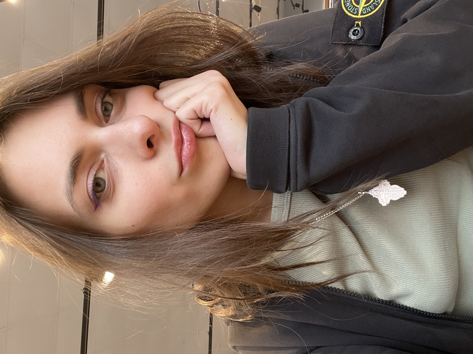 A young woman with light skin and green eyes gazes at the camera. Her brown hair is shoulder-length and parted. She wears light purple eyeliner on her lower lash line. Her lips are a pale pink shade. She is wearing a dark jacket with a Stone Island patch on the upper left arm and a light green sweater underneath. A silver necklace with an ornate pendant hangs around her neck. Her right hand is resting on her cheek, creating a slightly pensive expression.