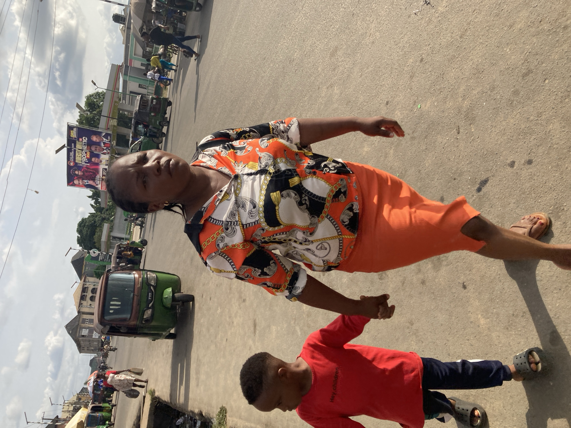 A woman and a child walk on a street in Aba, Nigeria. The woman wears a patterned shirt and orange skirt, with sandals on her feet. The child, presumed to be a young boy, wears a red shirt, navy pants, and Croc-style shoes. They are holding hands. The street is paved and has traffic, including motorcycles and three-wheeled vehicles. A billboard with various figures can be seen in the background. Buildings line the street, and the sky is partly cloudy.