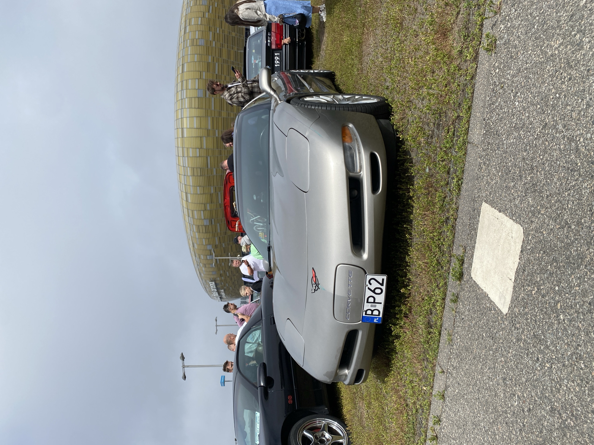 In Gdańsk, Poland, on an overcast day, a group of people gather near the PGE Arena. In the foreground, a silver Chevrolet Corvette C5 is parked on grass, with its front license plate reading "B P62". To the left, a black Chevrolet Camaro SS is partially visible, with the text "SOCHY-BOR" on its side. In the background, the distinctive golden, wave-like facade of the PGE Arena stands under a cloudy sky. Several individuals are milling about, some looking at the cars and others interacting with their phones. The scene appears to be an informal car show or gathering.