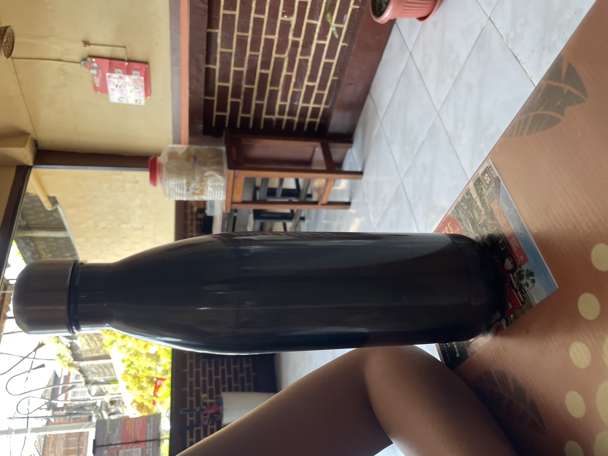 A large, dark brown insulated water bottle stands upright on a patterned table. To the left, a person's bare arm and shoulder are visible. In the background, a brick wall is present, and beyond that, a bright outdoor scene with greenery and buildings can be seen through a doorway. A wooden stool with a plastic container of white granular material sits on top of it. The floor is tiled. A red and white display rack with small items is mounted on the wall. The scene appears to be an indoor/outdoor cafe or restaurant setting in Denpasar, Indonesia.