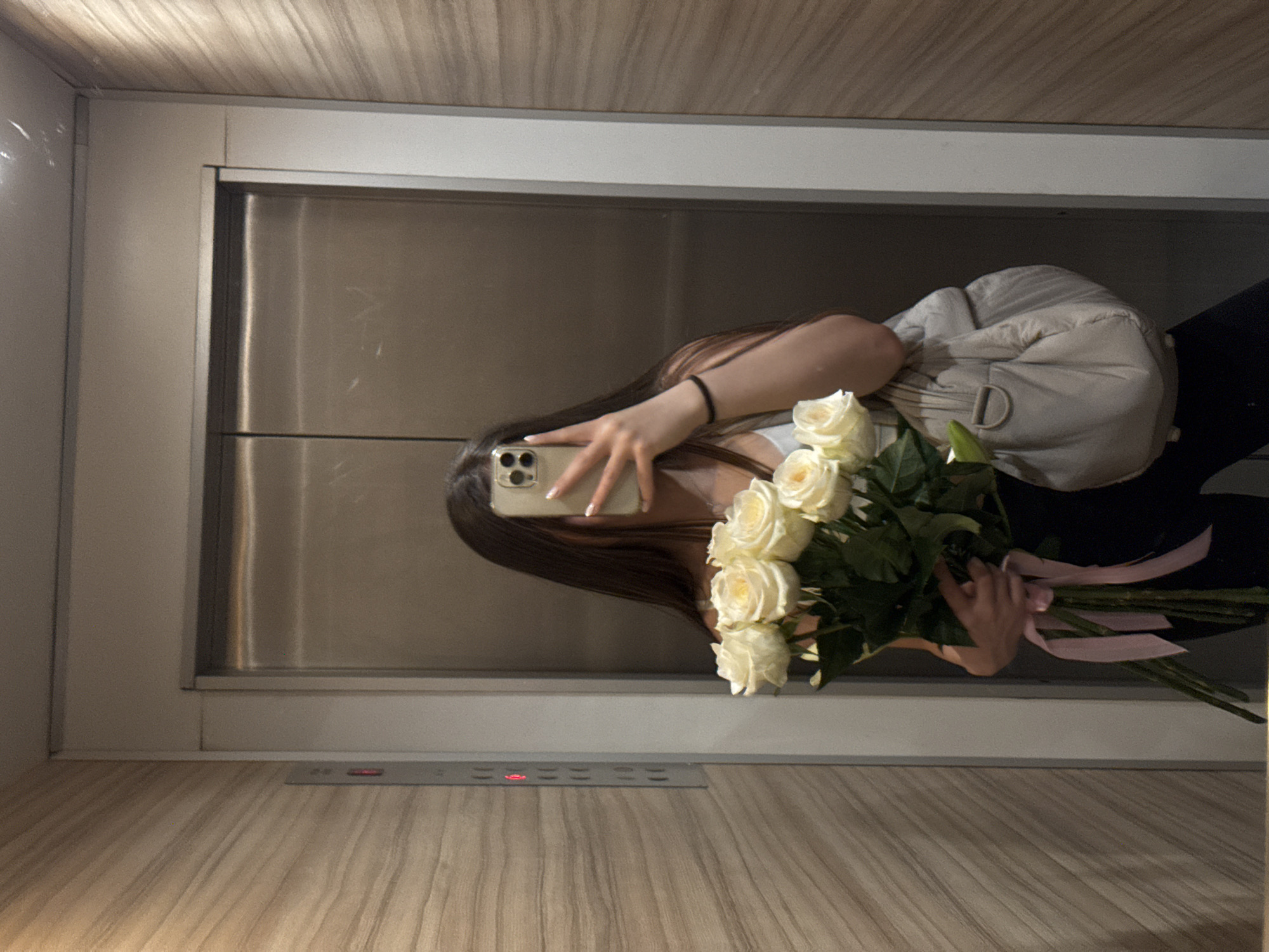A woman in an elevator is taking a photo of herself in a mirror. She is holding a bouquet of white roses tied with pink ribbons. The elevator has brushed metal doors and a wooden-patterned wall. The elevator buttons are visible. The city of Sofia, Bulgaria cannot be determined from the image.