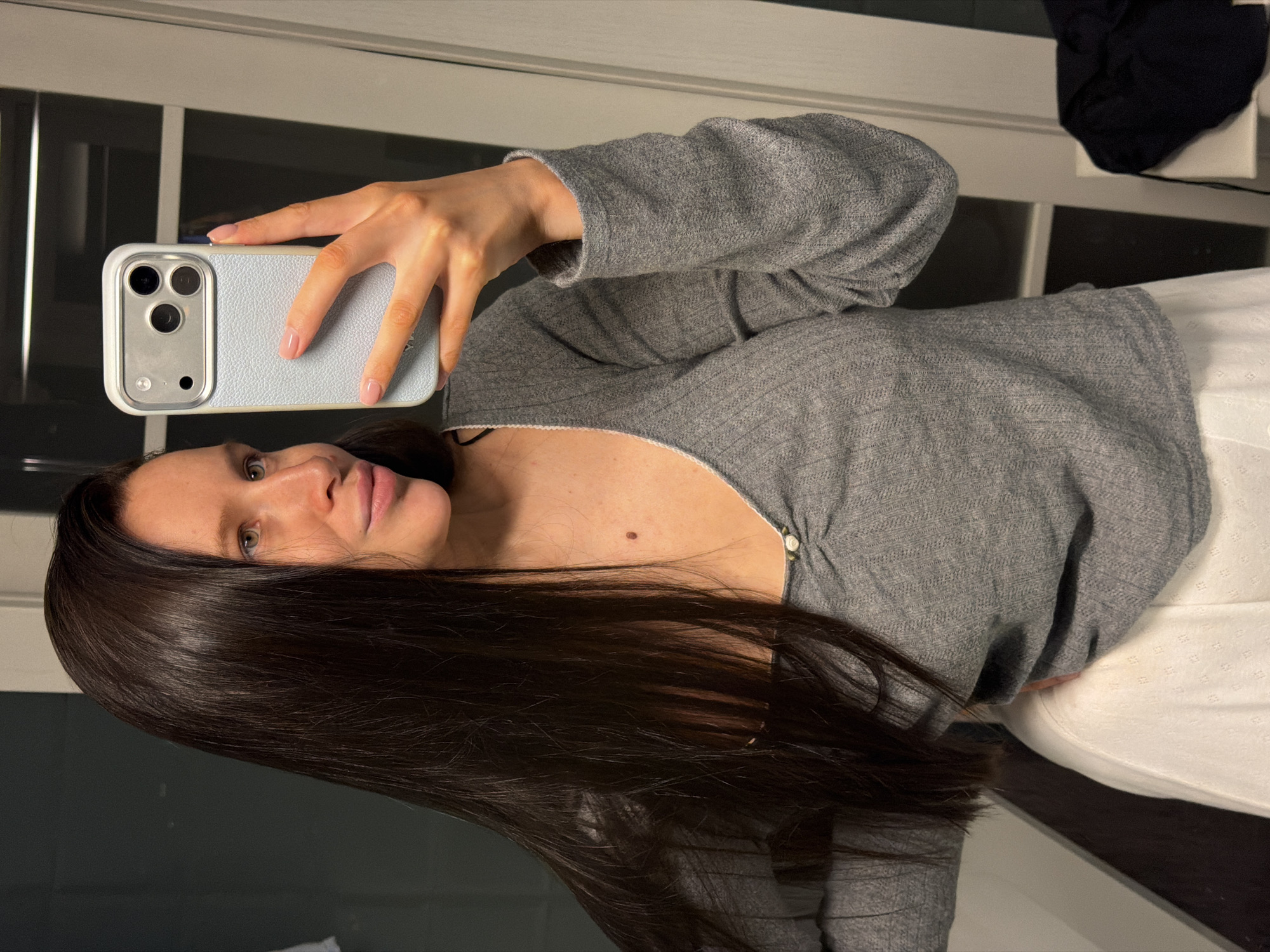 A young woman is taking a selfie in what appears to be a residential space with a window behind her. She is holding a light blue cell phone in her right hand. She is wearing a gray long-sleeved shirt and white bottoms. Her hair is long and dark brown. The location in the city of Khimki, Russia is not possible to determine from this image.
