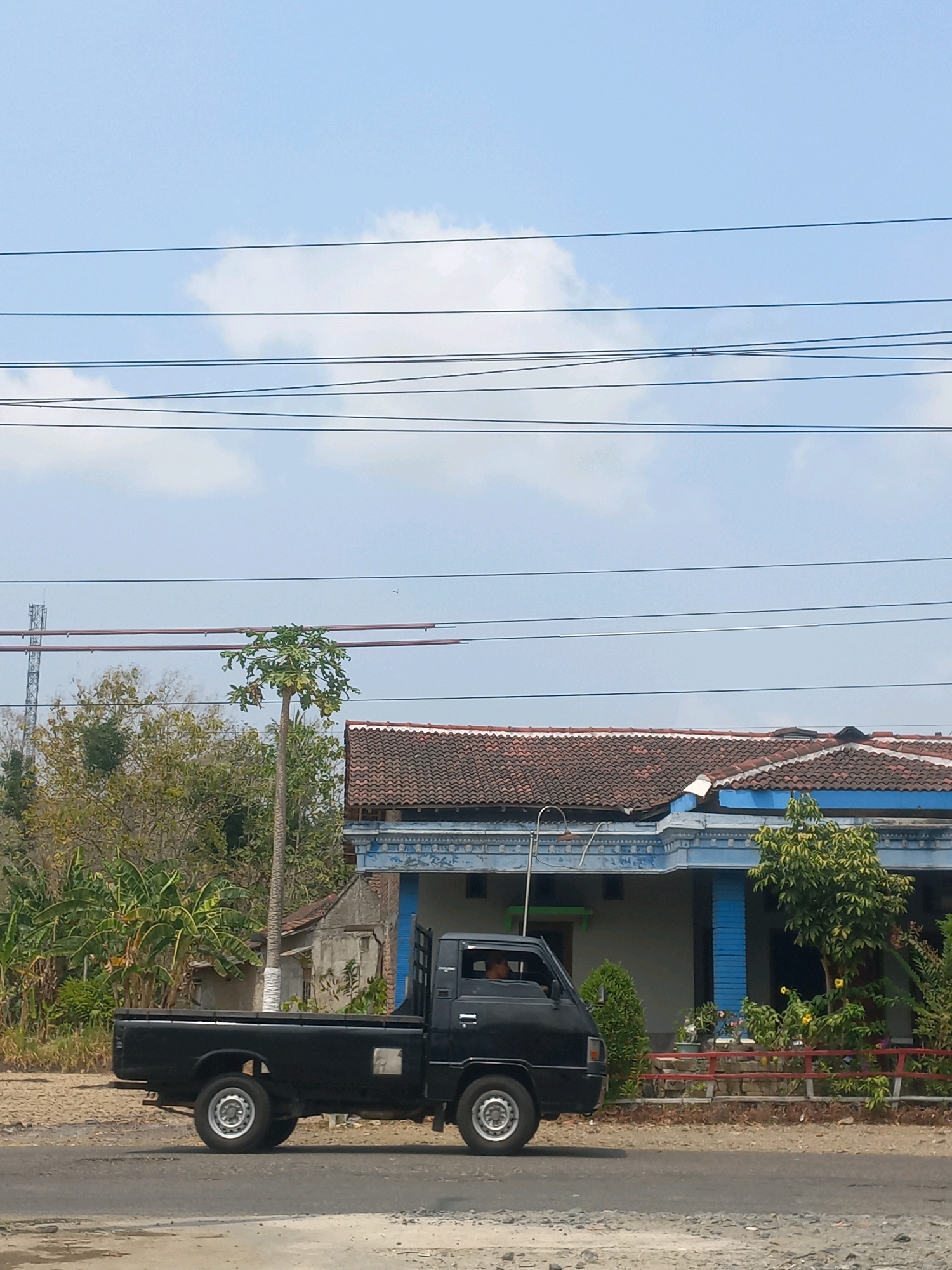 A black pickup truck drives on a paved road in front of a house in Baturetno, Indonesia. The house has a tiled roof and blue trim. There are several trees and plants around the house, and utility wires are strung across the sky above. The sky is blue with some white clouds. The truck has a driver visible through the windshield.