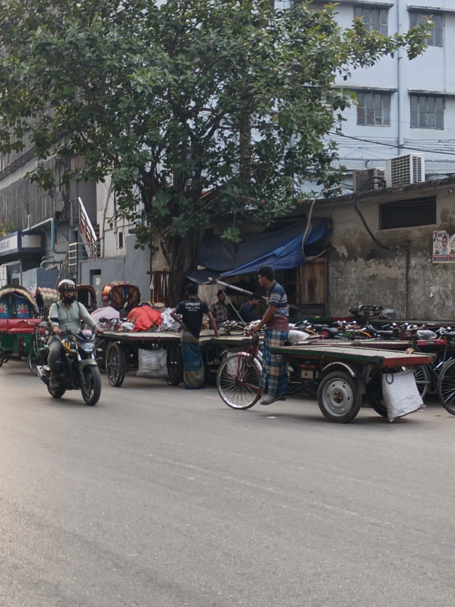 The image shows a street in Dhaka, Bangladesh. A man on a motorcycle drives along the road. Several carts are arranged along the side of the road. Some of the carts are topped with various goods. Several men stand around the carts. A large tree partially obscures buildings in the background. A row of parked bicycles sits behind the carts.