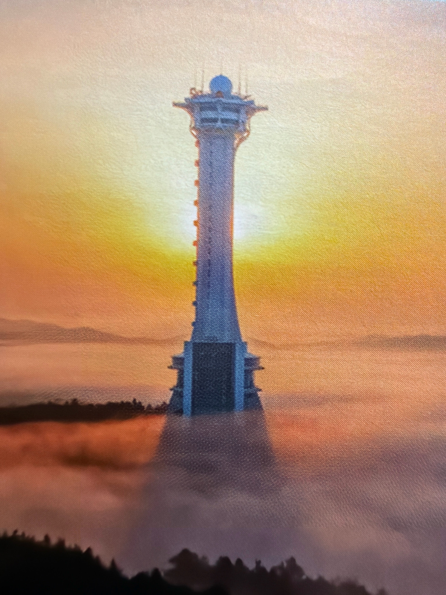 The image depicts a tall, slender tower with a distinctive silhouette, possibly a communication tower or lighthouse, bathed in the warm glow of a sunrise or sunset. The tower rises from a misty landscape, with layers of fog or clouds obscuring the ground below. Silhouetted trees and hills are visible in the foreground and middle ground, suggesting a natural or semi-rural setting. The sky is a vibrant gradient of yellow, orange, and pink, with the sun creating a bright halo effect behind the tower. The tower itself appears to be made of concrete or metal, with visible structural elements and antennae at the top. The bottom section of the tower has a wider, more block-like base. The overall atmosphere is serene and majestic. The location is identified as Hunchun, China, but no specific landmarks are discernible within the image itself to confirm this.