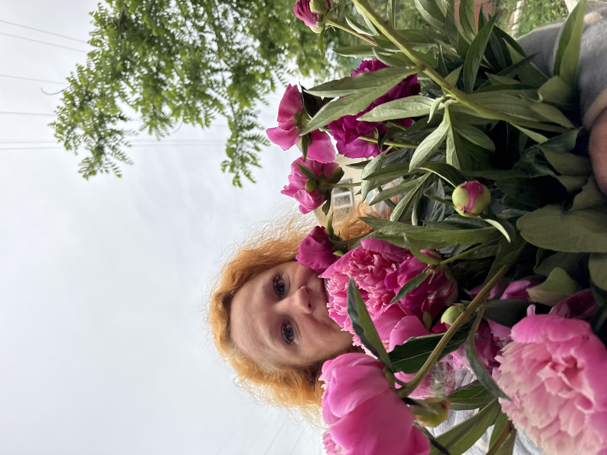 A medium shot photograph shows a woman with fiery red hair holding a bouquet of vibrant pink peonies in an outdoor setting. The woman's face is visible in the upper center of the image, with her gaze directed slightly off-camera. She is wearing a gray garment, and her hair is styled in a somewhat tousled manner. The peonies are the primary focus, with several large, fully bloomed flowers in the foreground, along with some buds. The background is blurred, revealing a pale, overcast sky, green foliage from a tree, and what appear to be utility wires. The overall scene suggests a casual, possibly spontaneous moment captured outdoors, in Chaikovsky, Russia.