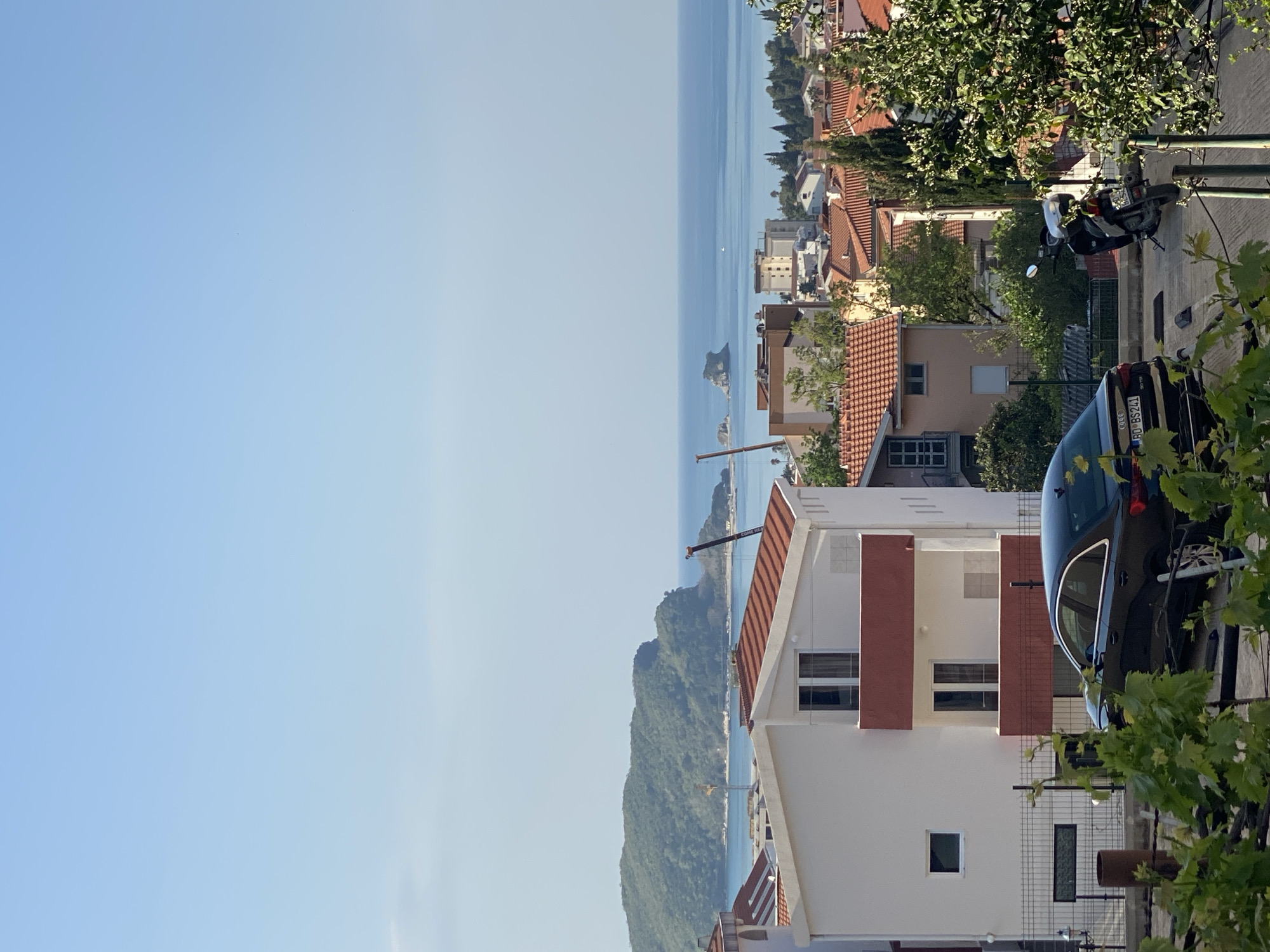 This image presents a panoramic, elevated view of a sunny coastal town, identified as Budva, Montenegro. The scene is captured on a clear day, indicated by the bright sunlight and a vast, light blue sky that dominates the upper portion of the frame.
In the middle ground, the calm, turquoise-blue waters of the Adriatic Sea stretch horizontally across the image. Several small, rocky islets or distant landmasses are visible in the sea, adding depth to the horizon. To the far left, a lush green, forested mountain slopes down to meet the coastline, with a construction crane also visible over the water in that area.
The lower and right sections of the image are filled with residential buildings featuring traditional red-tiled roofs, typical of Mediterranean architecture. These structures are nestled amidst abundant green foliage and trees, suggesting a verdant environment.
In the immediate foreground on the right, a prominent multi-story white building with reddish-brown accented balconies and windows is visible. A black Audi car is parked on the right, partially obscured by vibrant green vine leaves, with its license plate clearly displaying "BD-BS241," which confirms the location as Budva. Further back on the right, a motorcycle is also parked near other buildings, and a vertical street lamp post stands nearby.
The overall impression is one of a peaceful, picturesque coastal settlement bathed in natural daylight, with no discernible human activity but conveying the serene ambiance of a bright, clear day.